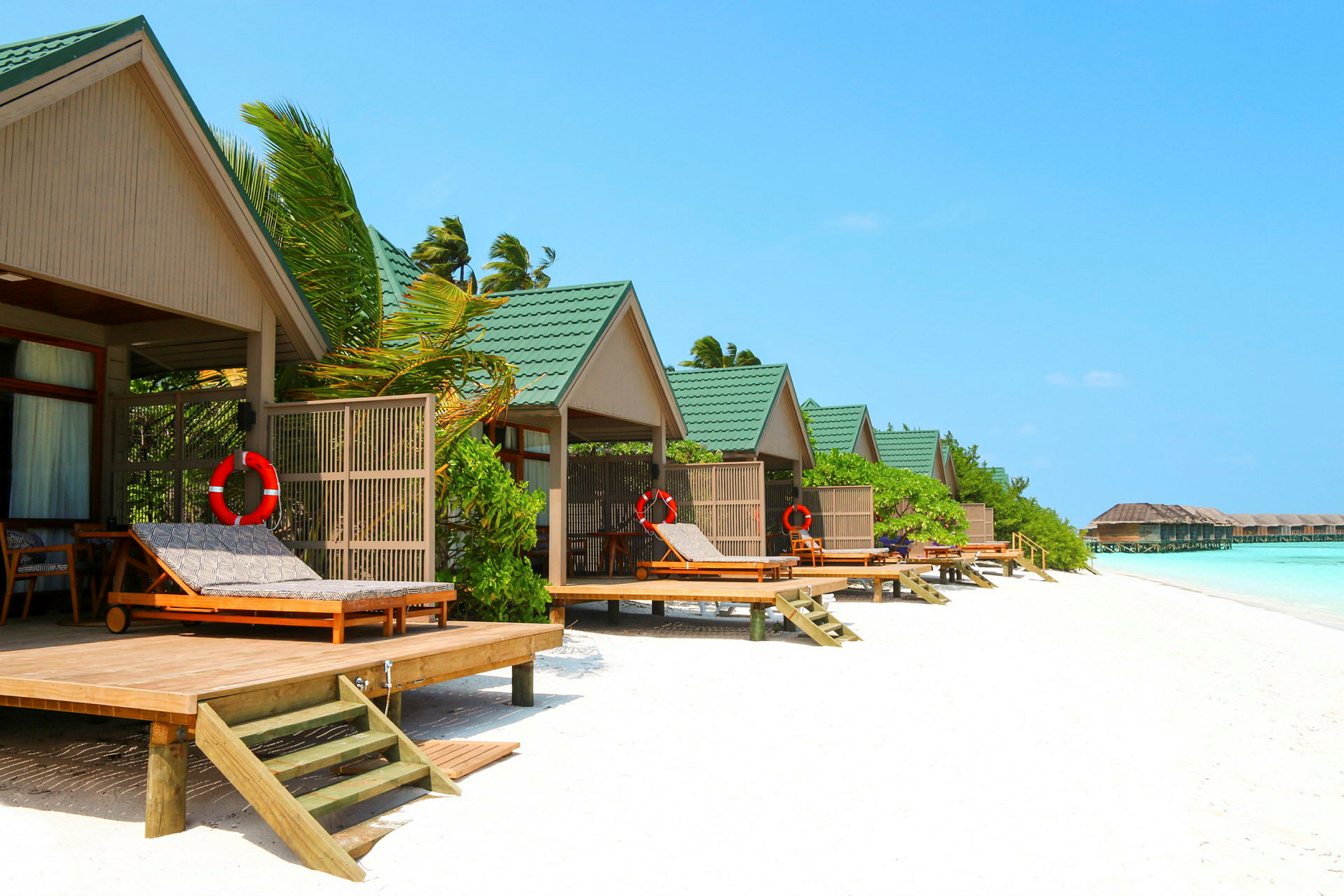 Strandbungalower med grön tak och solstolar vid vattnet. (AI-genererad text)