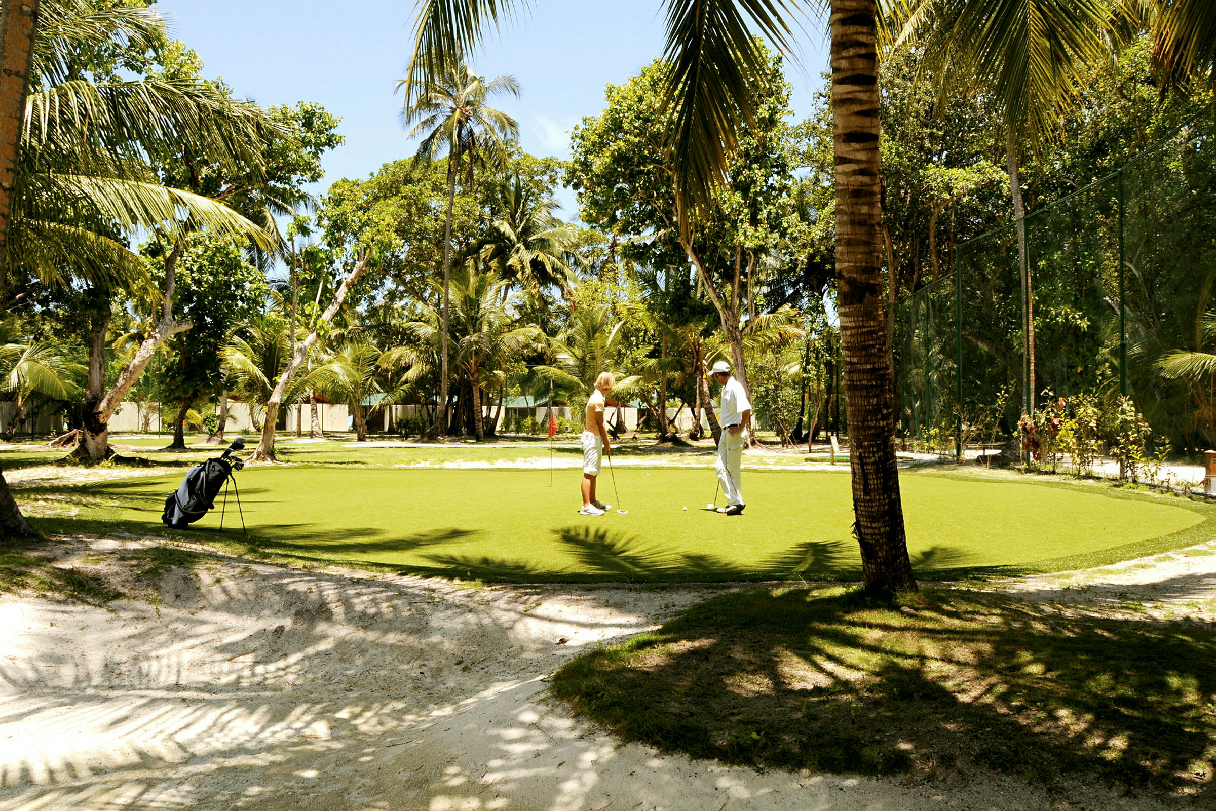 Två personer spelar golf på en grön puttinggreen omgiven av palmer. (AI-genererad text)