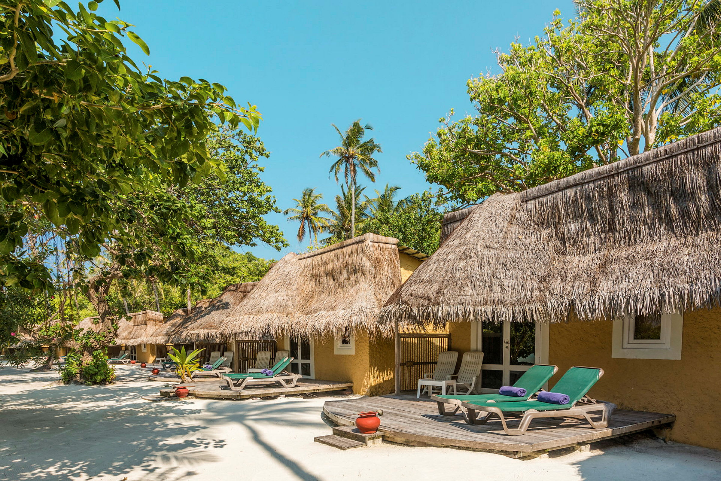 Strandbungalow med halmtak och solstolar framför omgiven av grönska. (AI-genererad text)