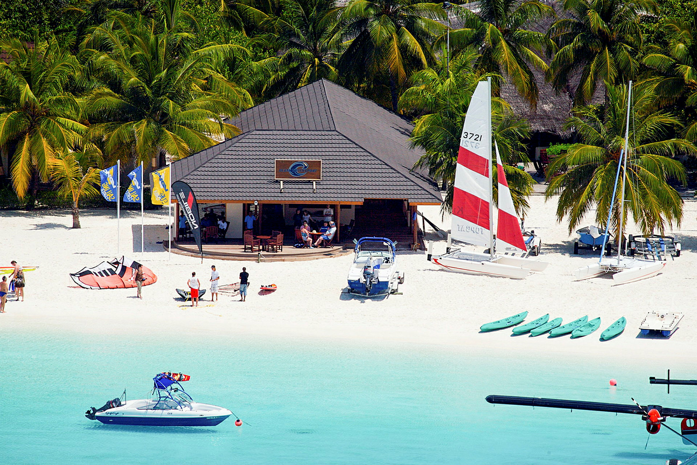 En strand med en båtklubb, katamaraner, kajaker och människor vid strandkanten. (AI-genererad text)