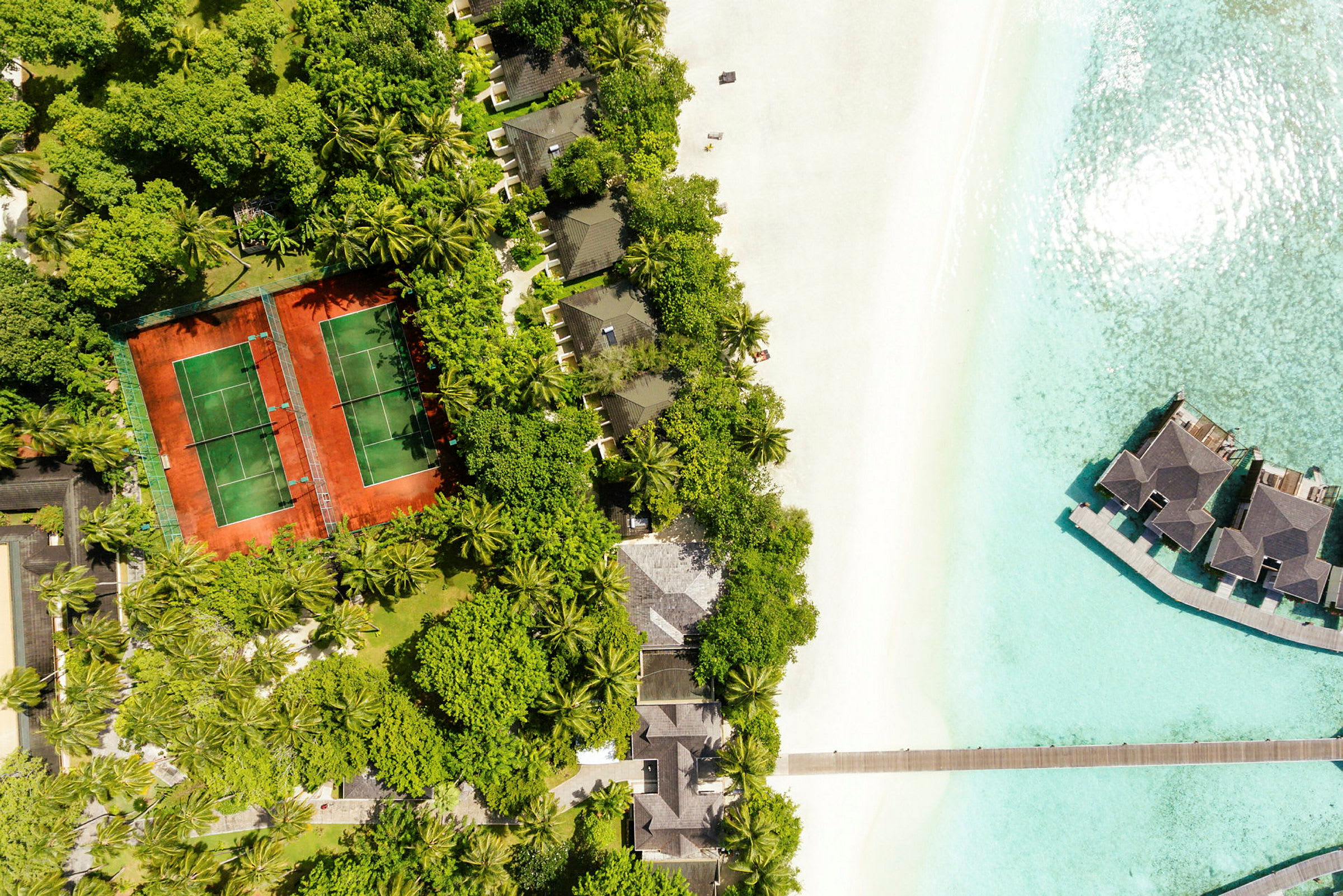Flygbild av en tropisk resort vid stranden med tennisbanor, villor bland träd och övervattensbungalower. (AI-genererad text)