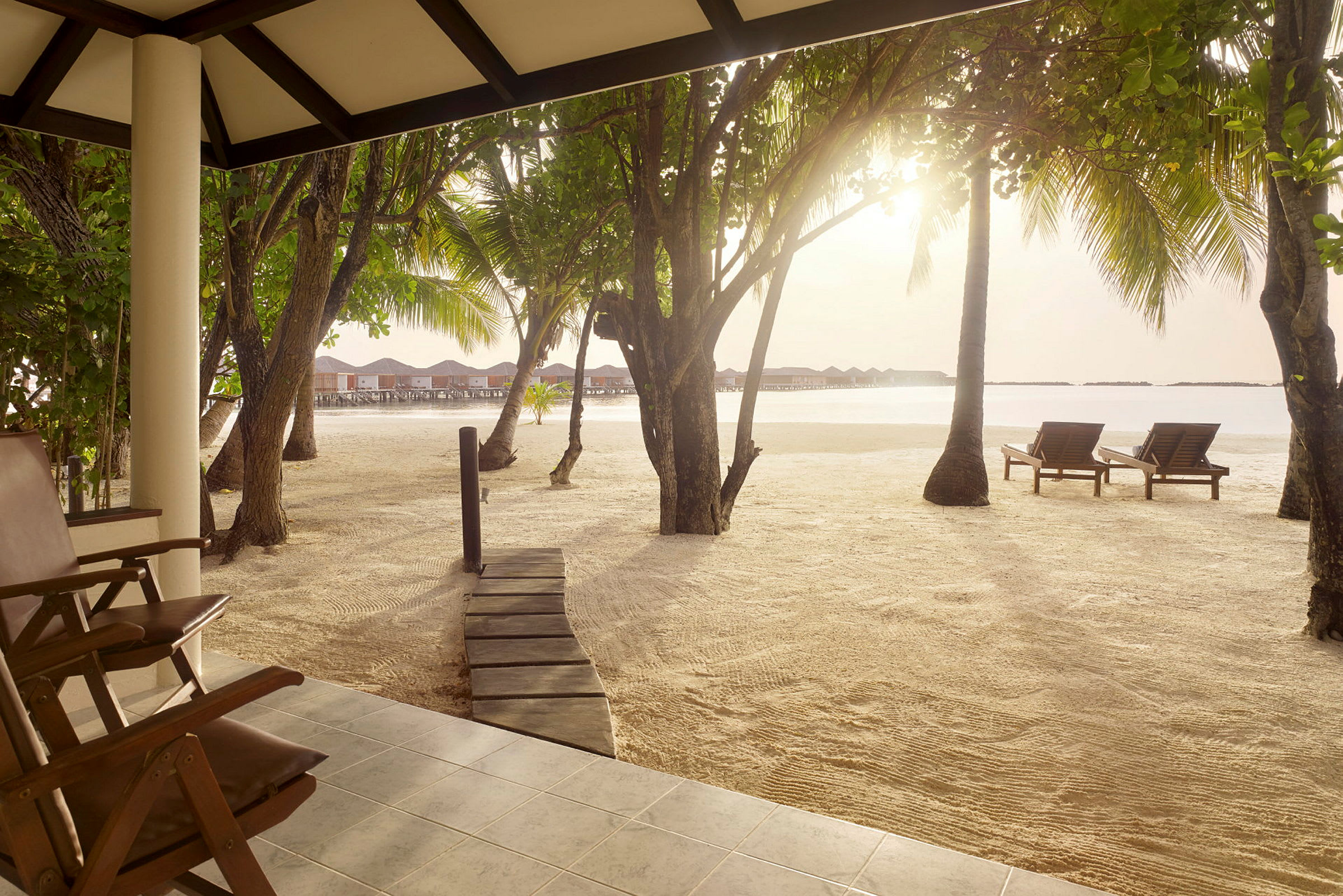 Vy från en terrass mot en sandstrand med solstolar, träd och bungalows på vattnet i bakgrunden. (AI-genererad text)
