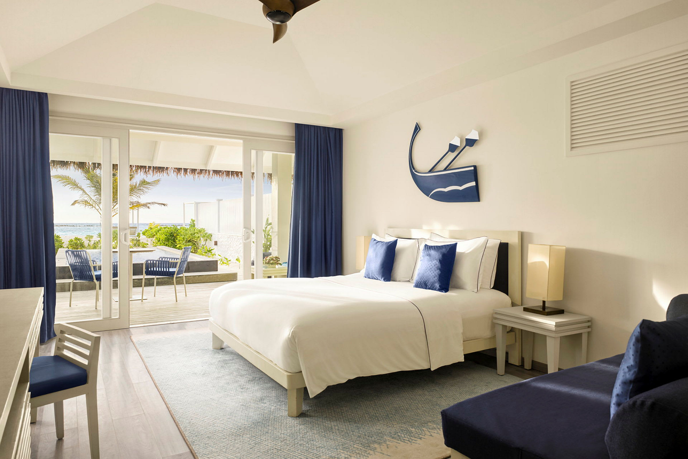 Ett hotellrum med en stor säng, blåa gardiner och vita möbler. Utsikt över en terrass och grönska utanför. (AI-genererad text)
