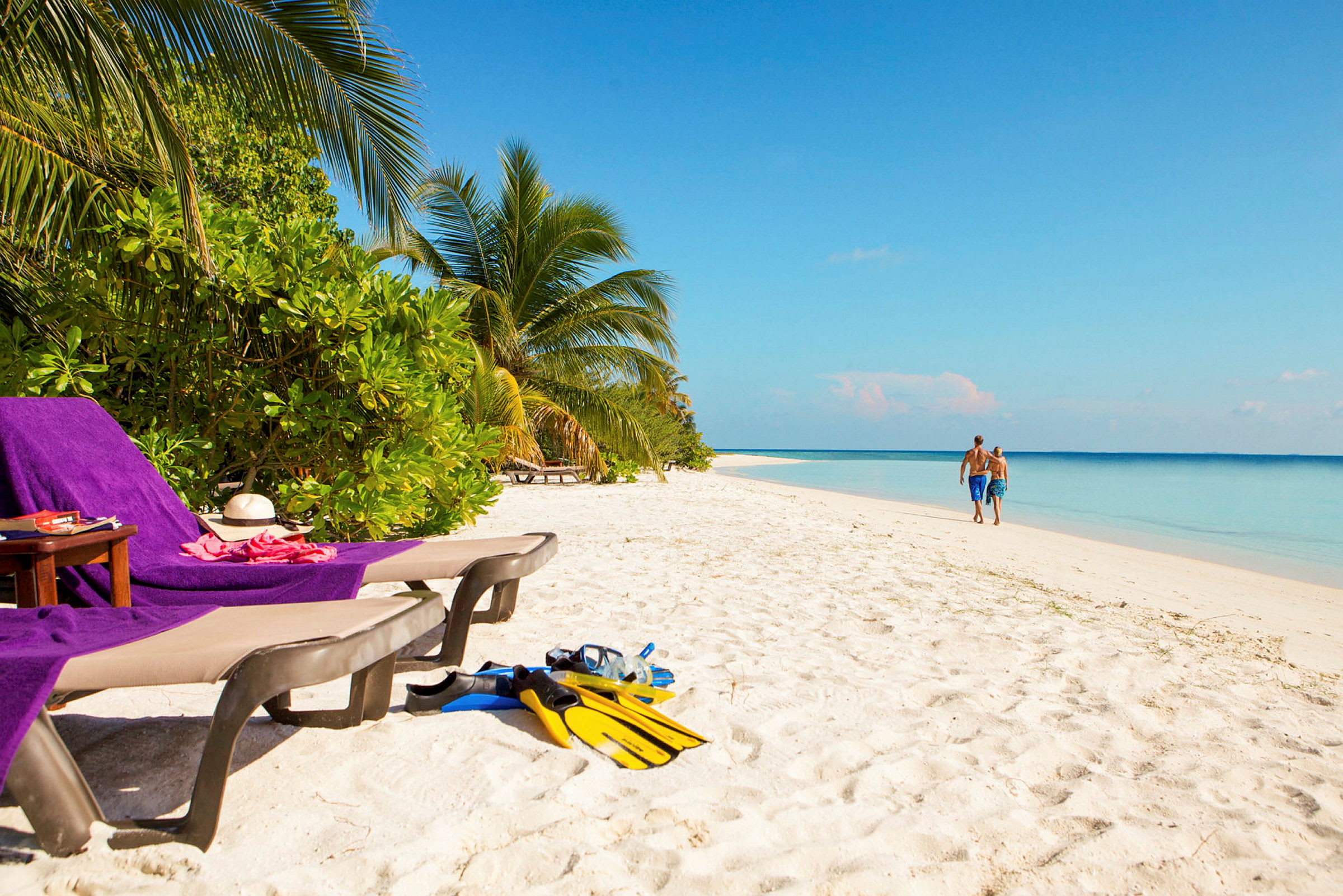 Sandstranden med solsängar, handdukar och snorkelutrustning. Två personer promenerar vid vattenkanten. (AI-genererad text)