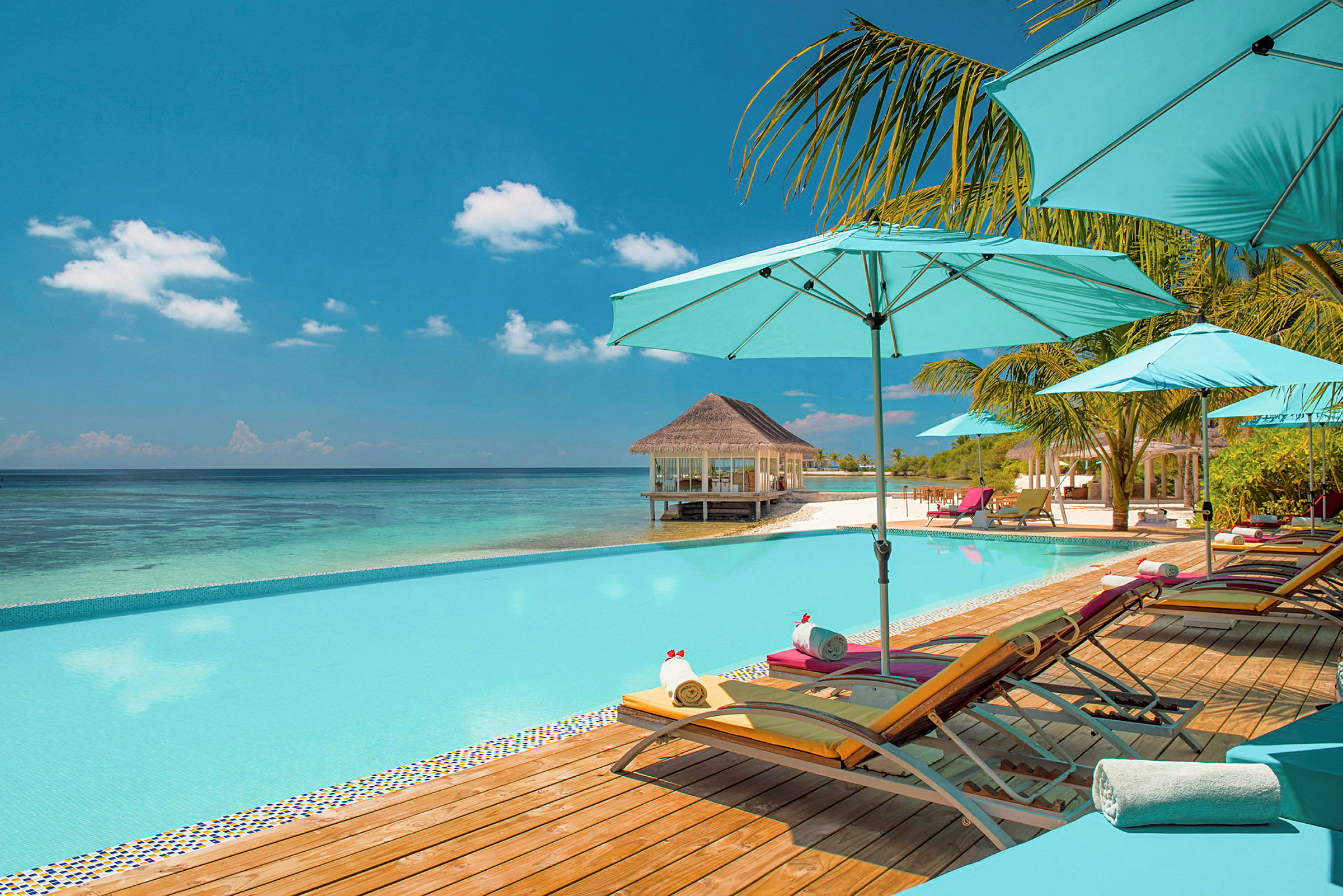 Pool med solsängar och parasoller vid havet, med en strand och en bungalow i bakgrunden. (AI-genererad text)