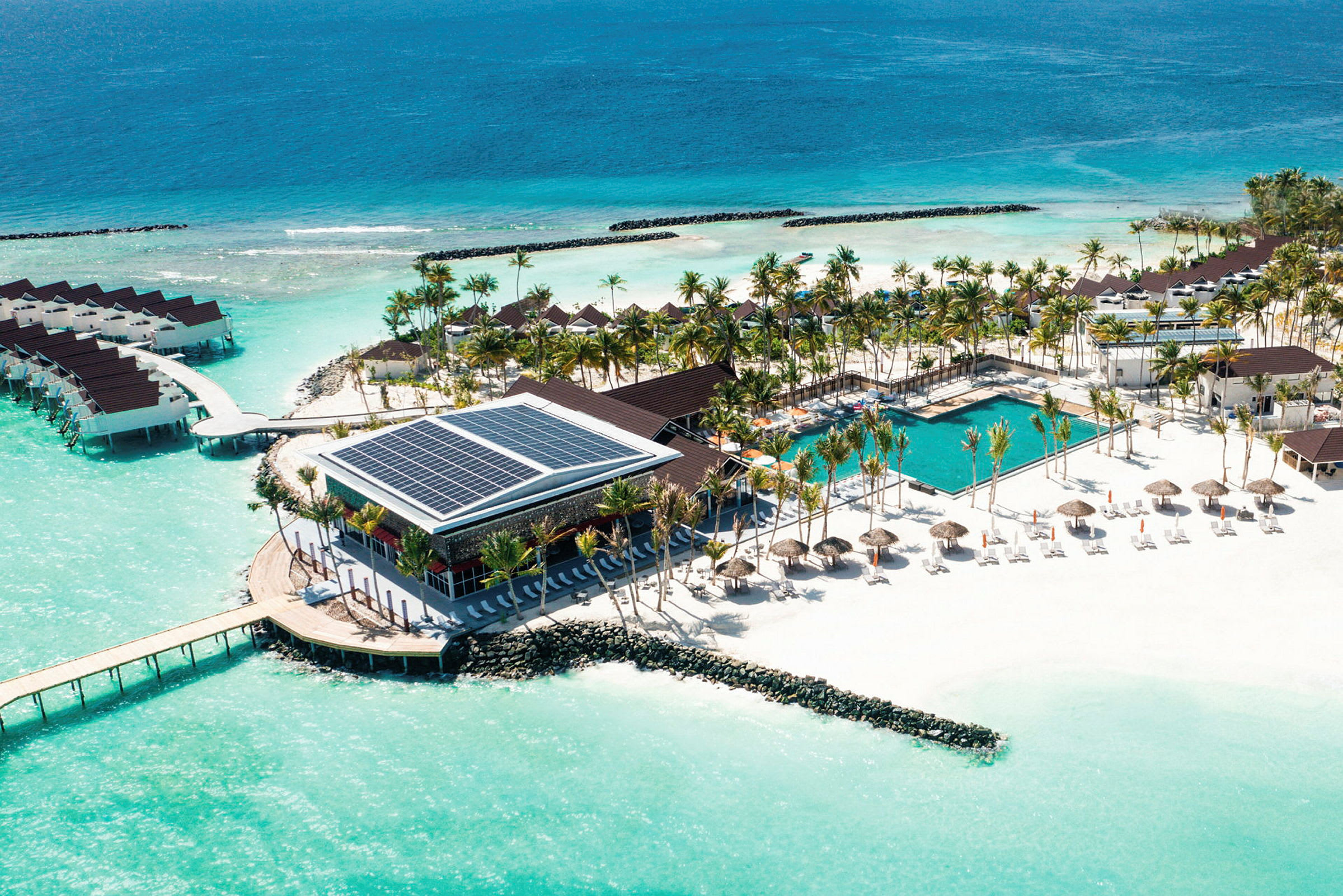 Flygfoto av en resort vid stranden med pool, solstolar och bungalows på vattnet. (AI-genererad text)