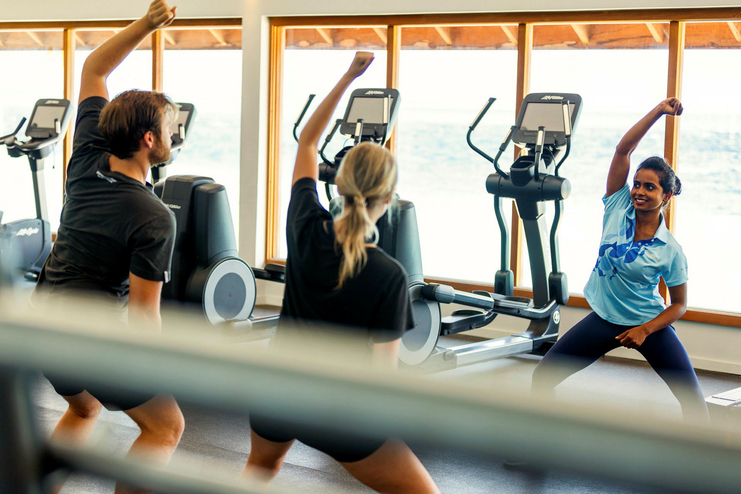 Personer utför träningsövningar i ett gym med konditionsmaskiner och stort fönster mot havet. (AI-genererad text)