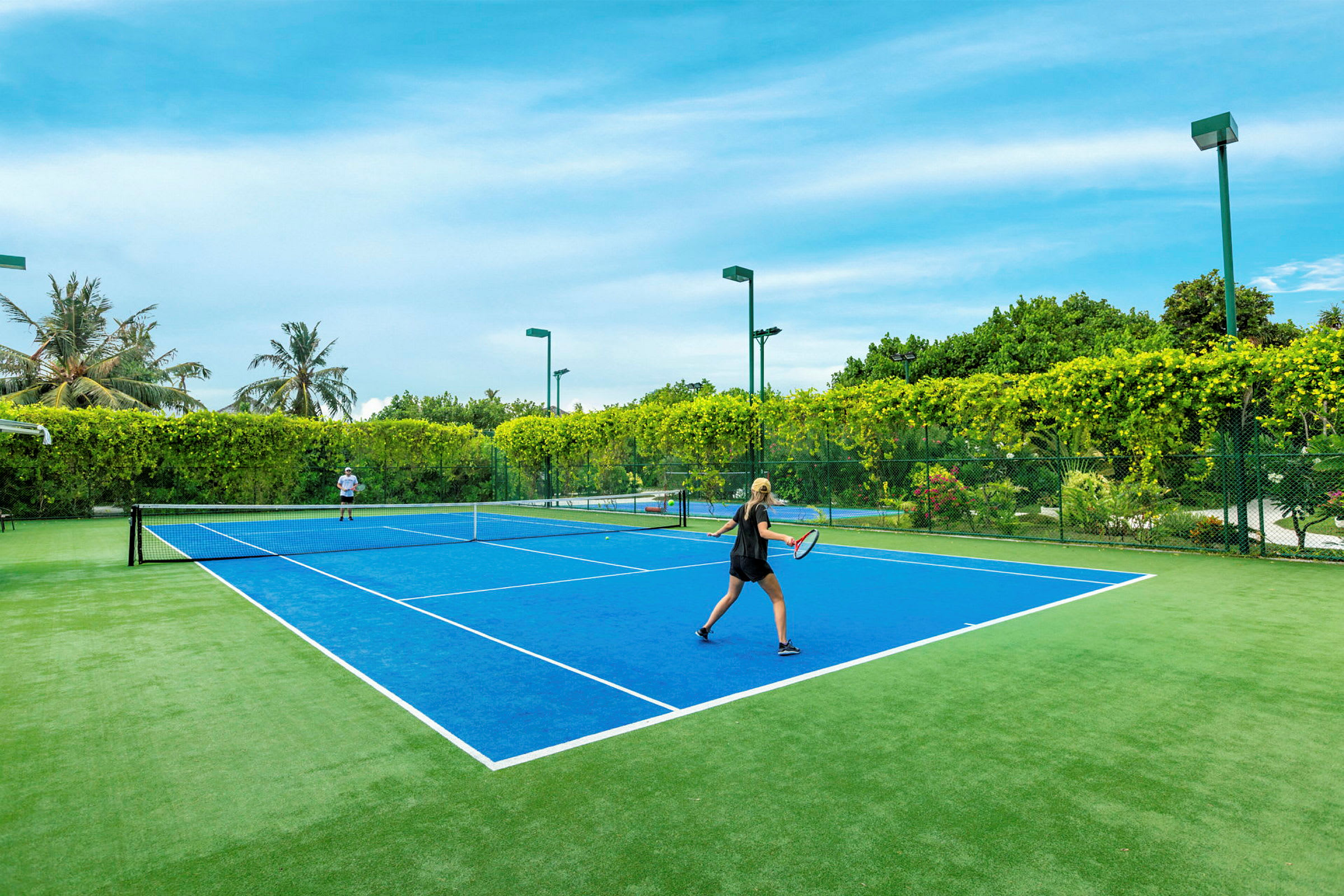 Två personer spelar tennis på en blå tennisbana omgiven av grönska. (AI-genererad text)