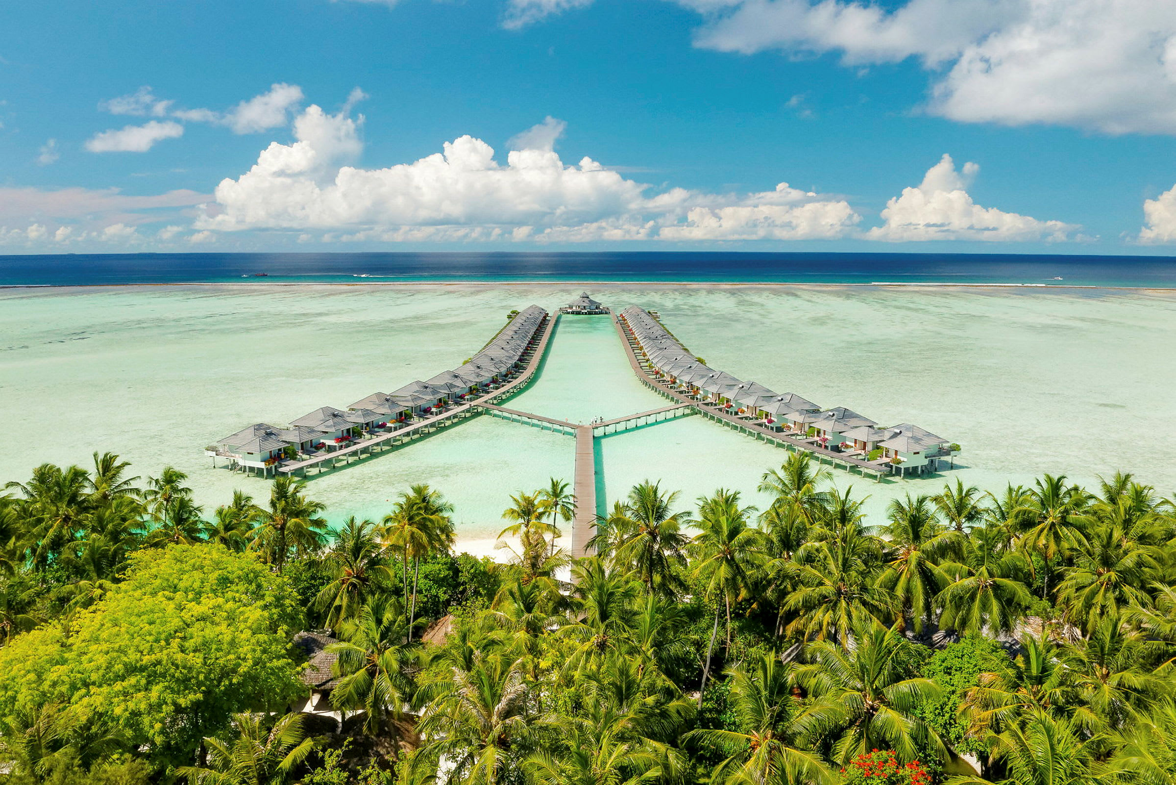 Bild av övervattenbungalower anslutna med bryggor över en lagun, omgivna av palmer och omgiven av blå himmel och hav. (AI-genererad text)