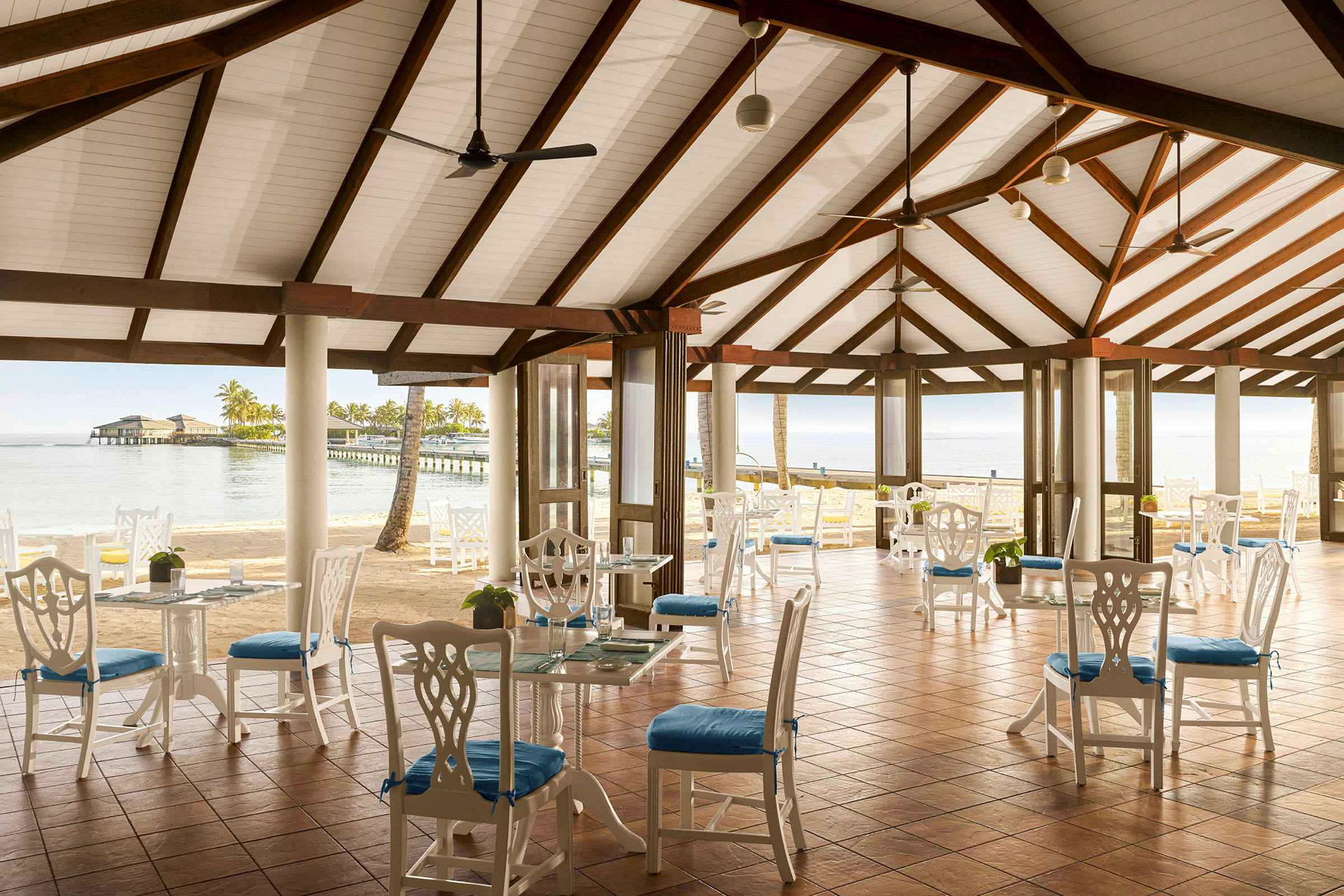 Bild av en öppen restauranglokal med vitmålade möbler och blå sittdynor, med utsikt över havet och en brygga. (AI-genererad text)