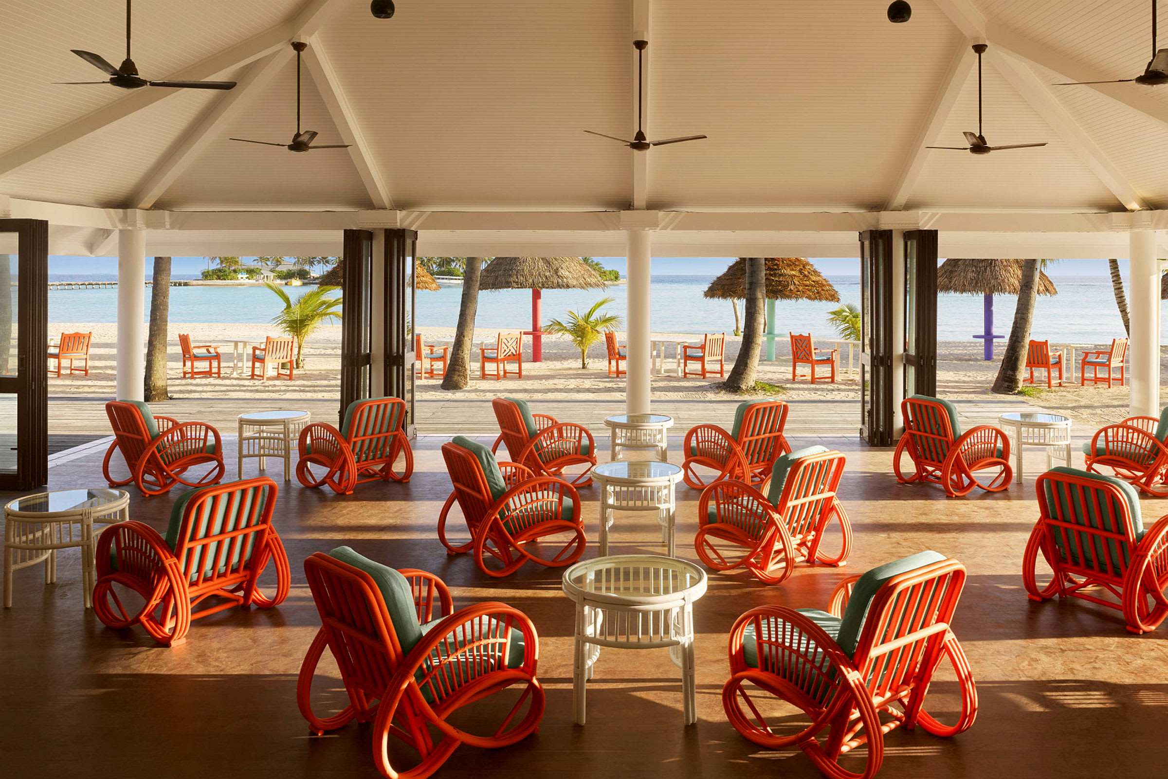 En öppen lounge med färgglada rottingstolar och bord, med utsikt över stranden och havet. (AI-genererad text)