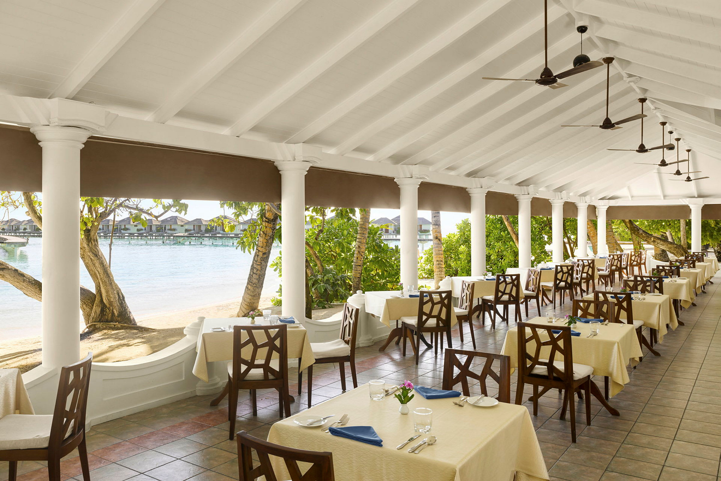 En restaurangs uteplats med dukade bord, omgiven av pelare och utsikt över stranden och vattnet. (AI-genererad text)