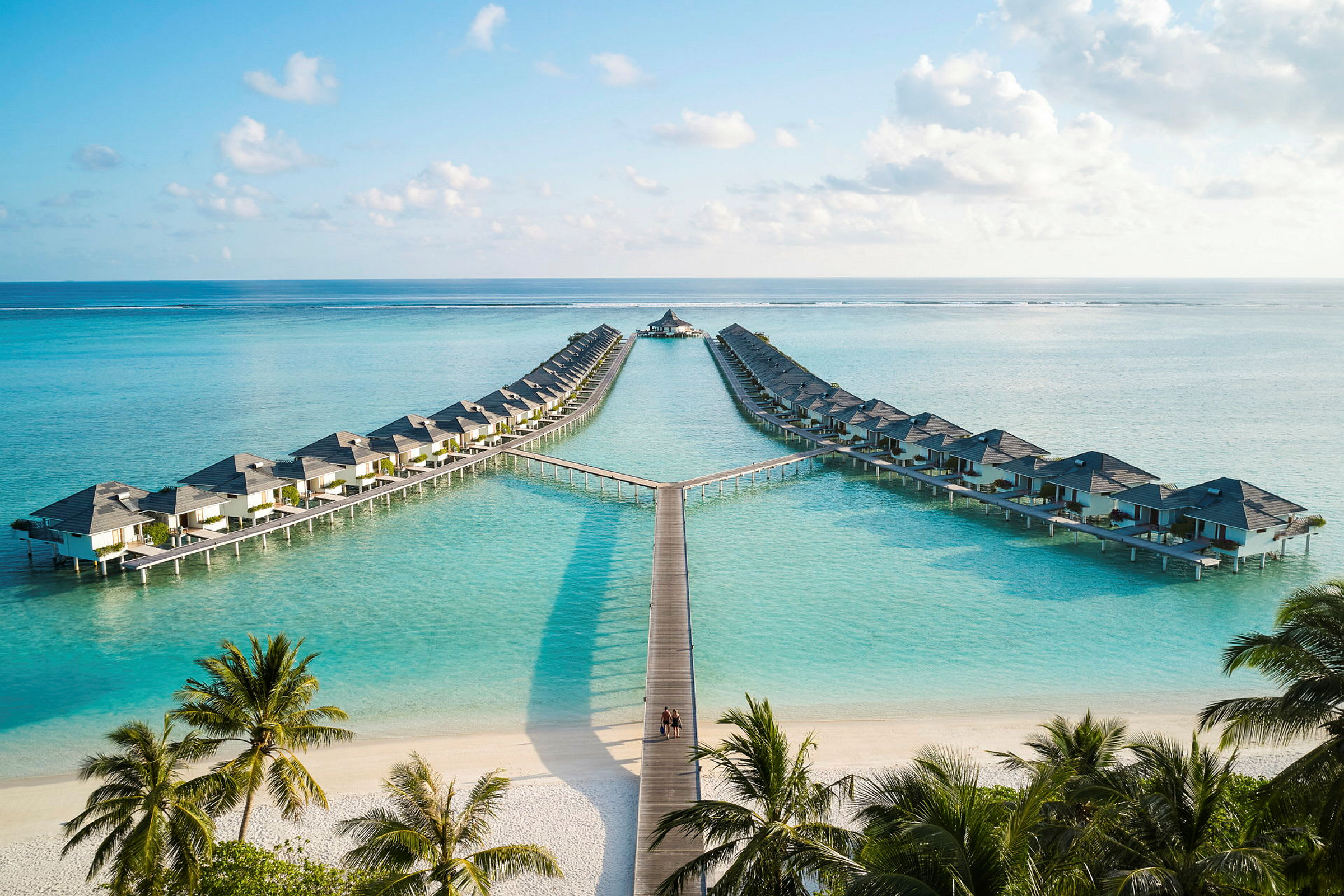 Hotellbungalower på pålar över klart blått vatten, med sandstrand och palmträd i förgrunden. (AI-genererad text)
