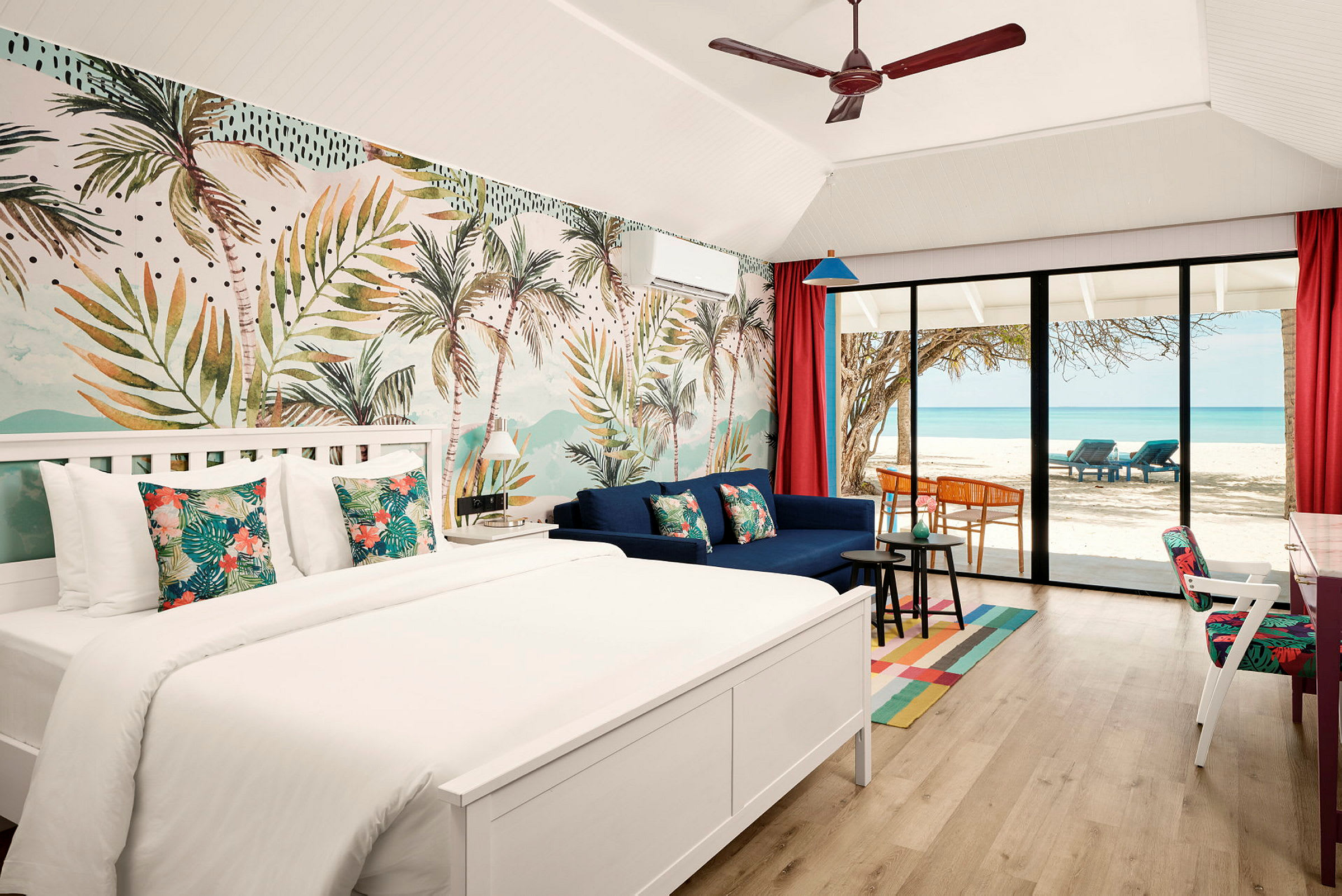 Ett hotellrum med en dubbelsäng, soffa och en stor glasdörr som vetter mot en strand med palmer och havsutsikt. (AI-genererad text)