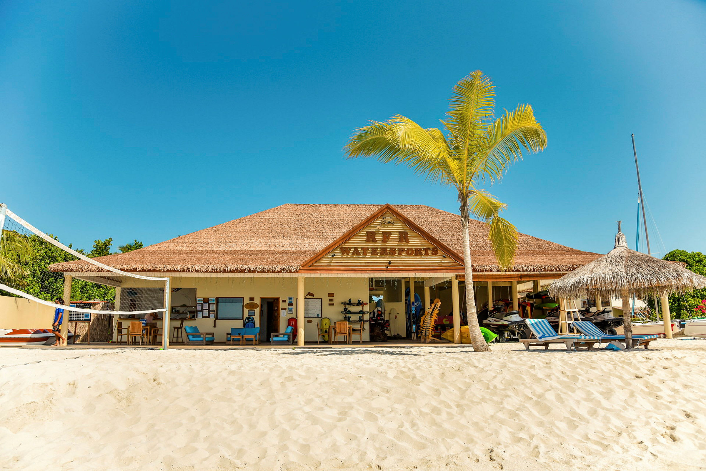 En byggnad med skylt "Reef Waterports" på en strand, med solstolar och en volleybollplan. (AI-genererad text)