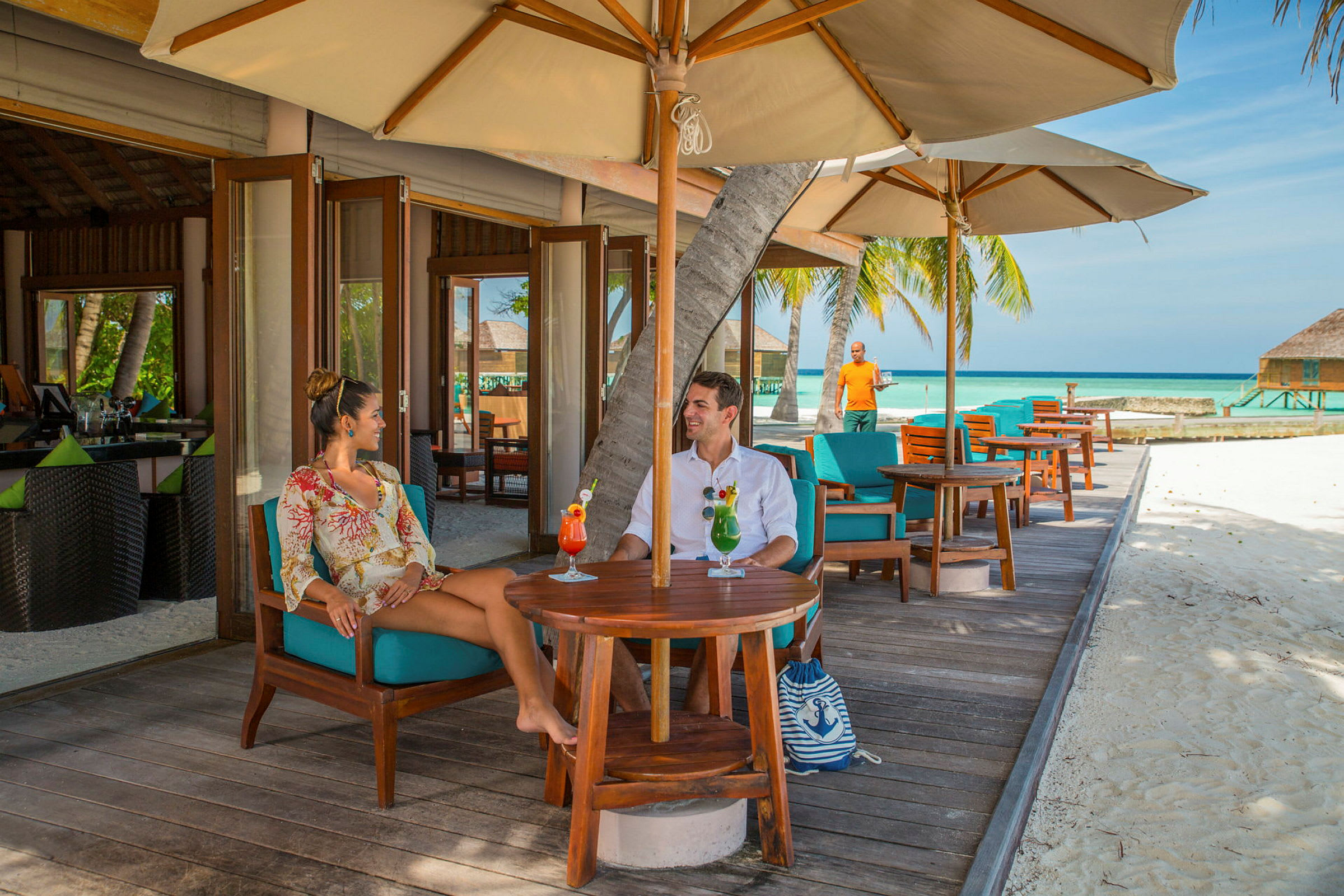 Två personer sitter vid ett bord på en utomhusrestaurang med utsikt över sandstrand och hav. (AI-genererad text)