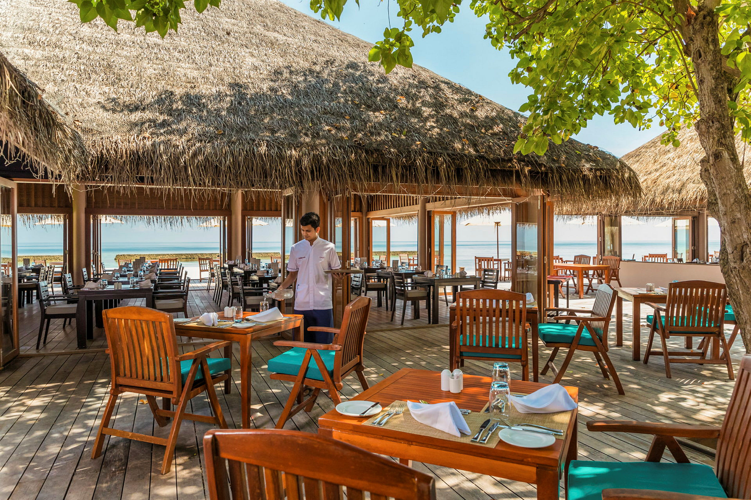 En utomhusrestaurang med bord och stolar av trä, halmtak och utsikt över havet. (AI-genererad text)