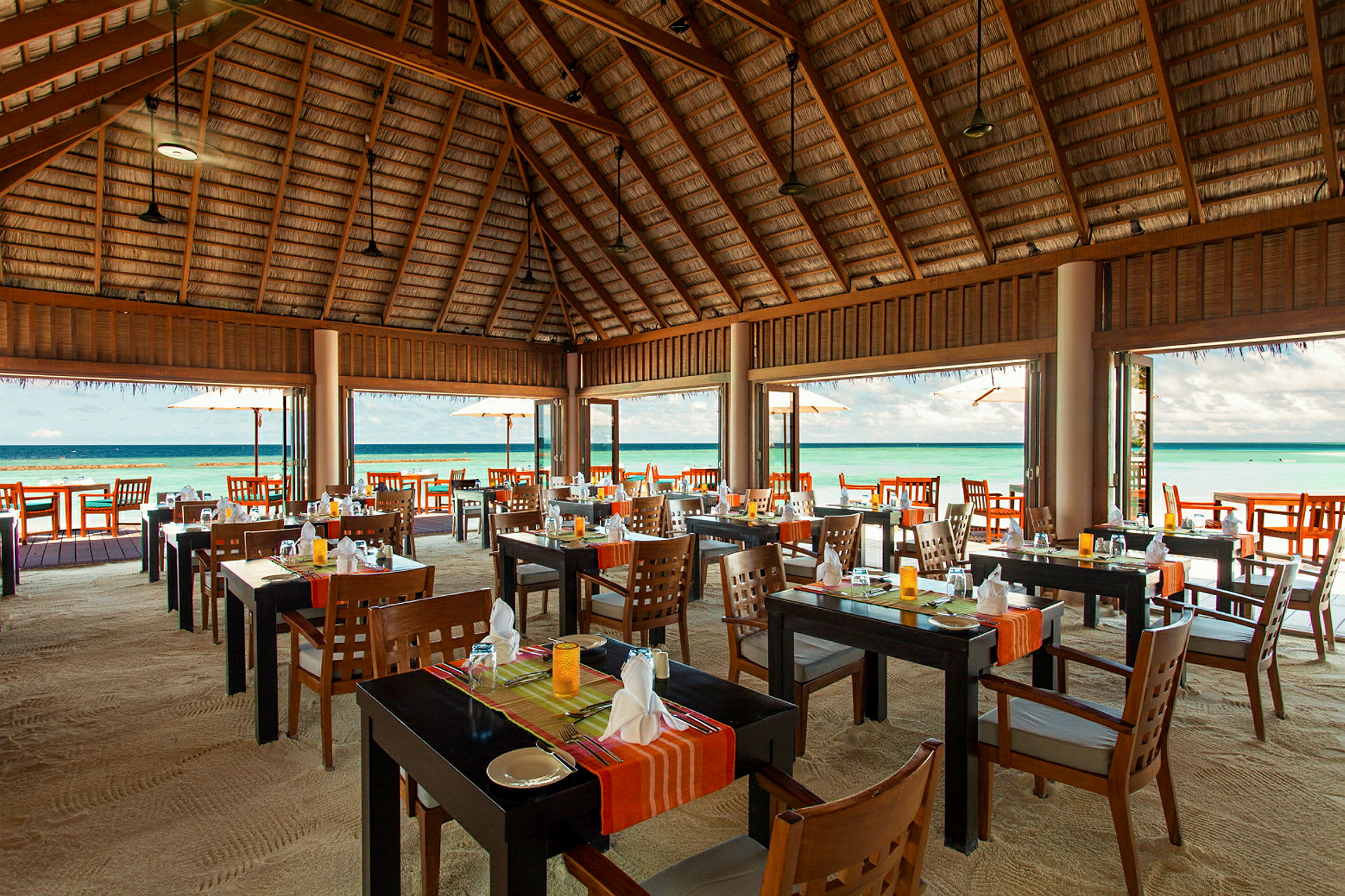 Restaurang med öppet tak av trä och bord på sand nära havet. (AI-genererad text)