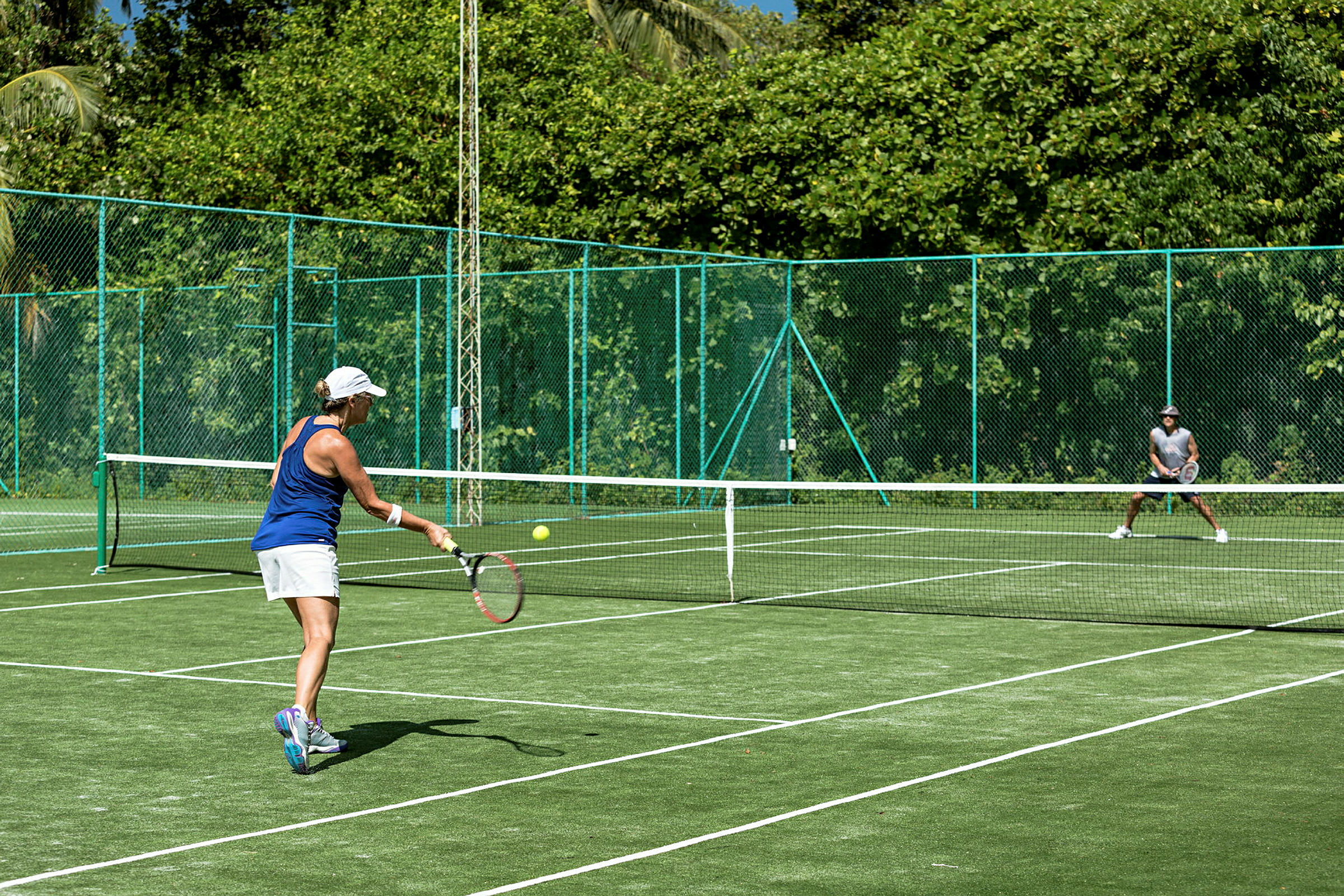 En person spelar tennis på en grön tennisplan omgiven av ett grönskande område. (AI-genererad text)