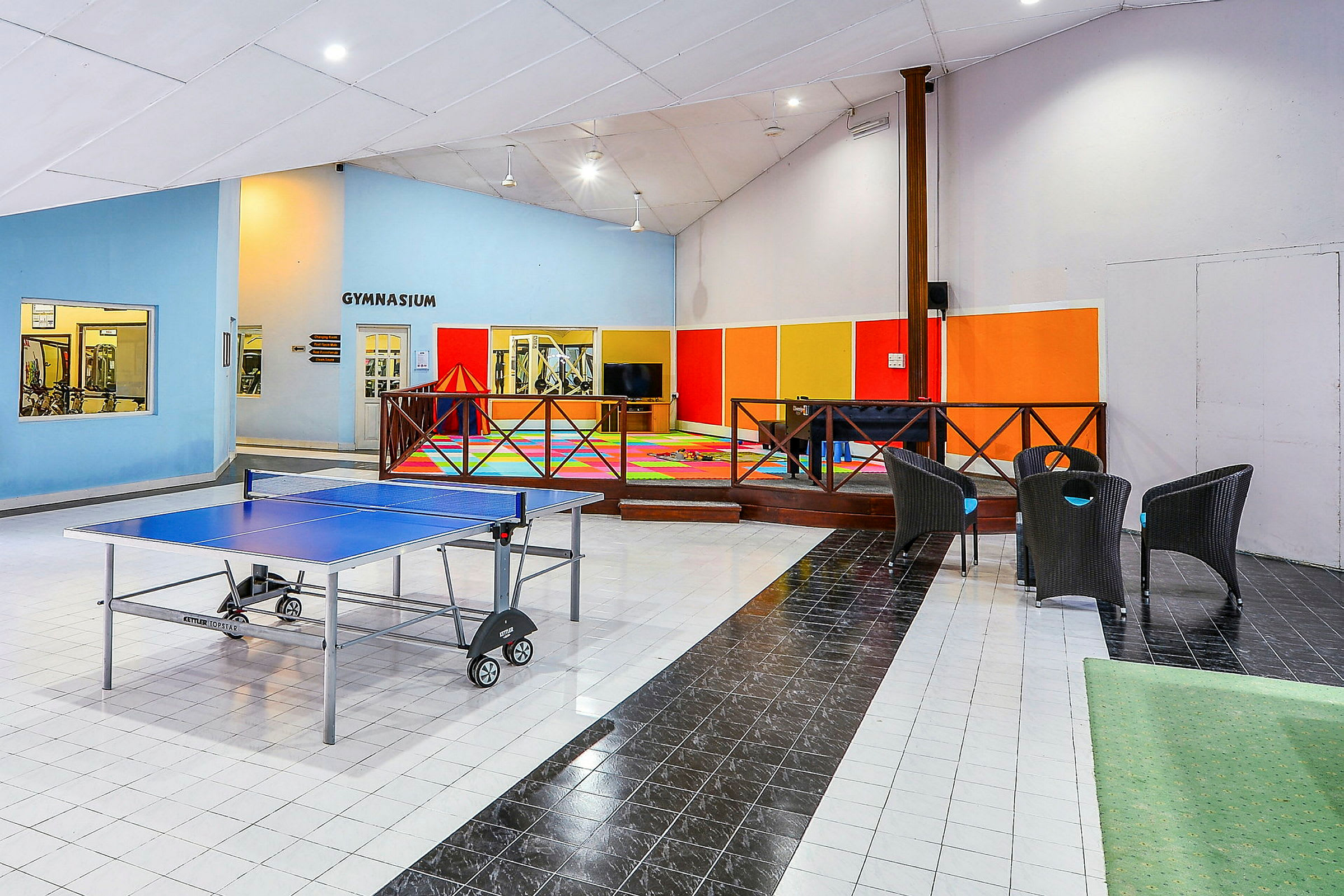 Bild av ett inomhus gemenskapsområde med ett pingisbord, sittplatser och färgglad lekhörna, samt en intilliggande gym-del. (AI-genererad text)