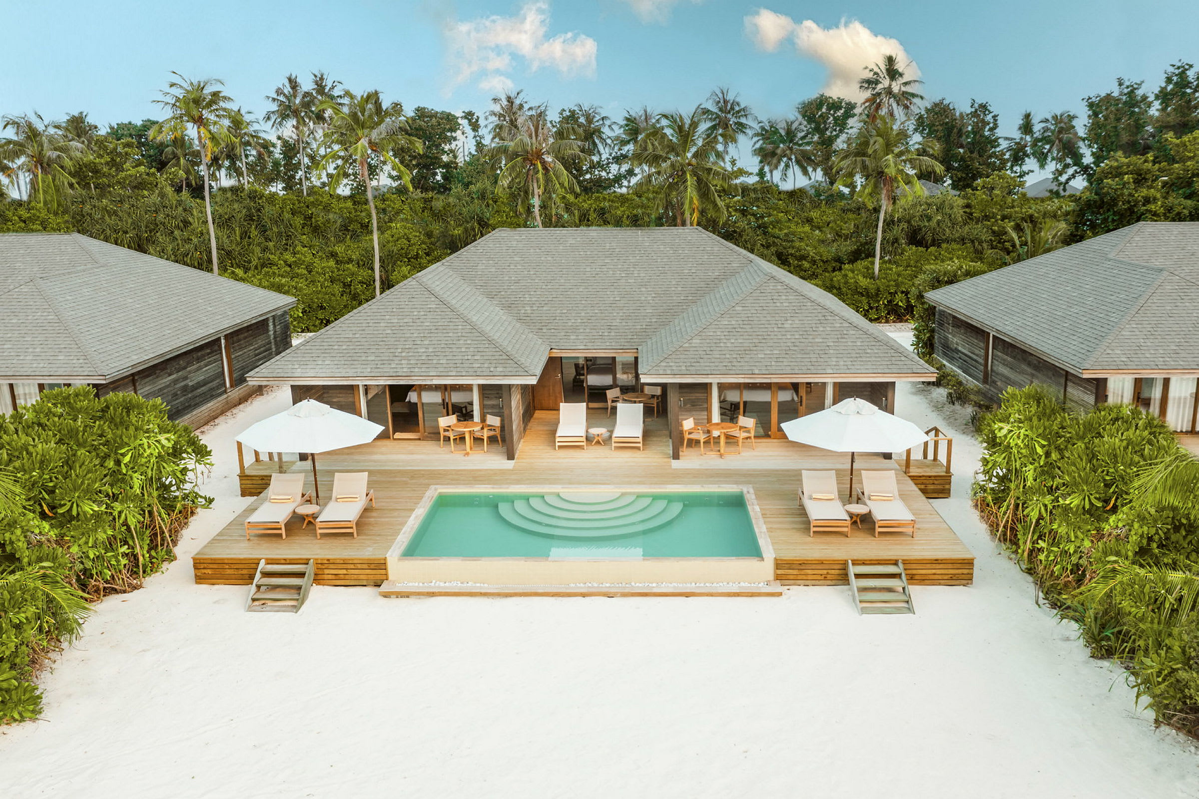 Villor med privata pooler och solterrasser omgivna av grönska vid en vit sandstrand. (AI-genererad text)