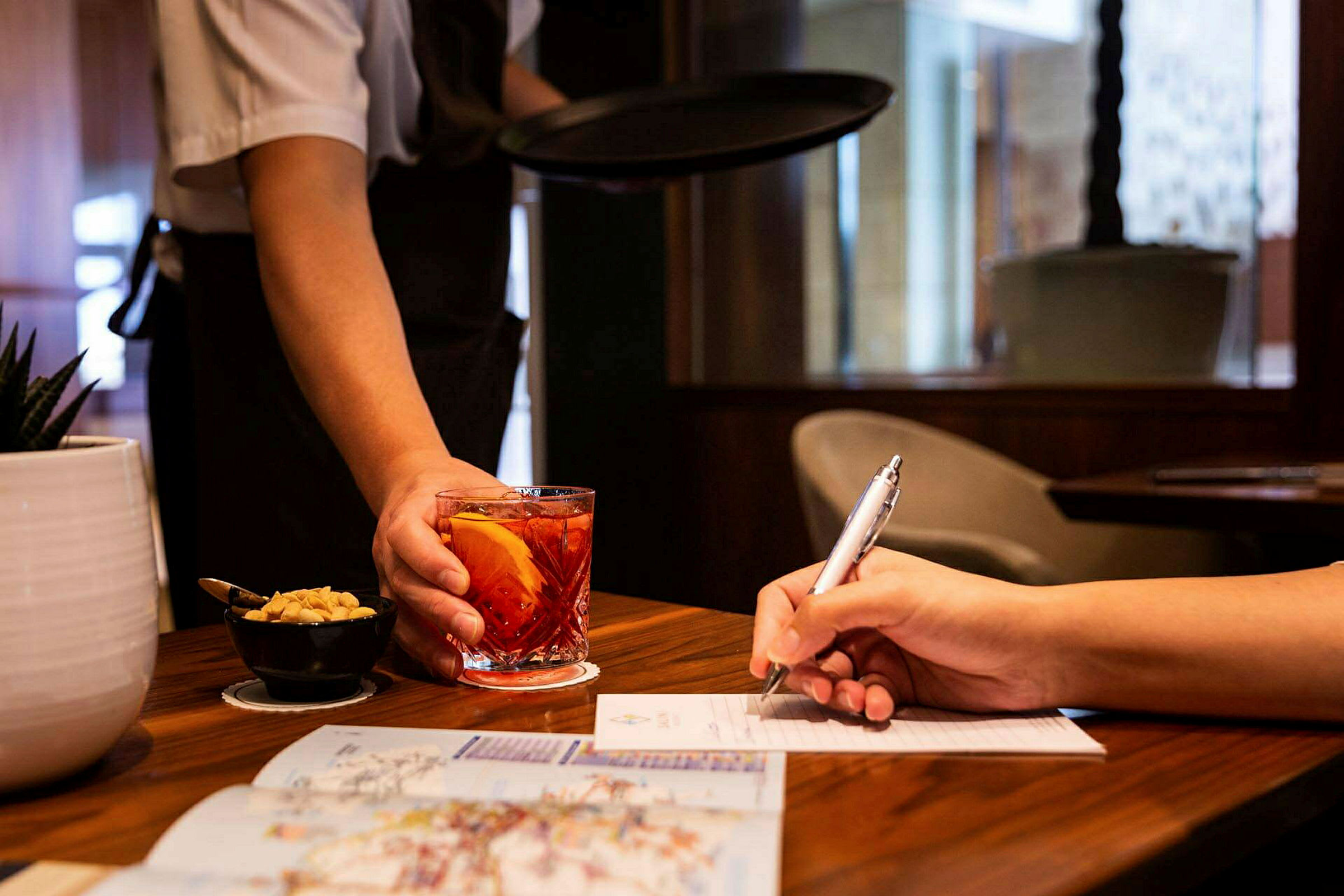 En servitör serverar en dryck i ett glas bredvid en person som skriver på ett papper. (AI-genererad text)