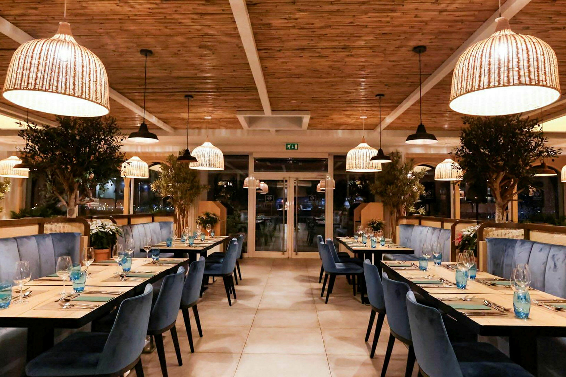 En elegant restauranginteriör med blå stolar, träbord och stora växtdekorationer. Ljuskronor i rottingstil hänger från taket. (AI-genererad text)