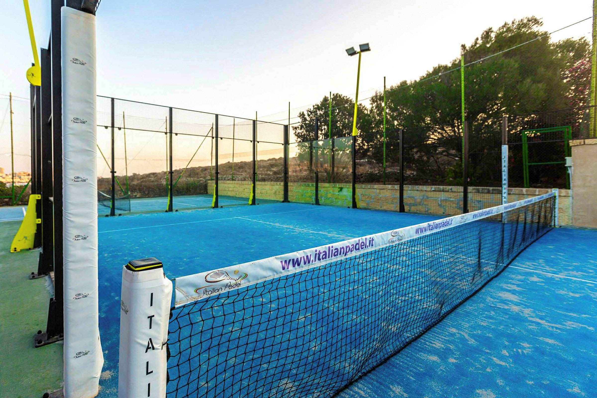 En padelbana med blå markeringar, omgiven av nät och stängsel i en utomhusmiljö. (AI-genererad text)