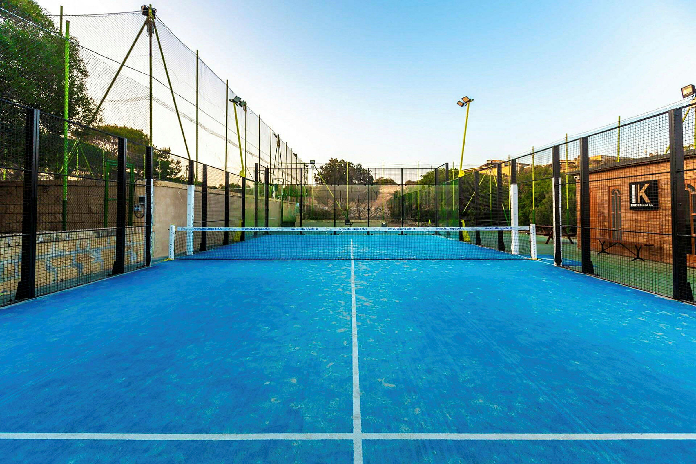 En padelbana med blå yta och nät i mitten, omgiven av stängsel. (AI-genererad text)