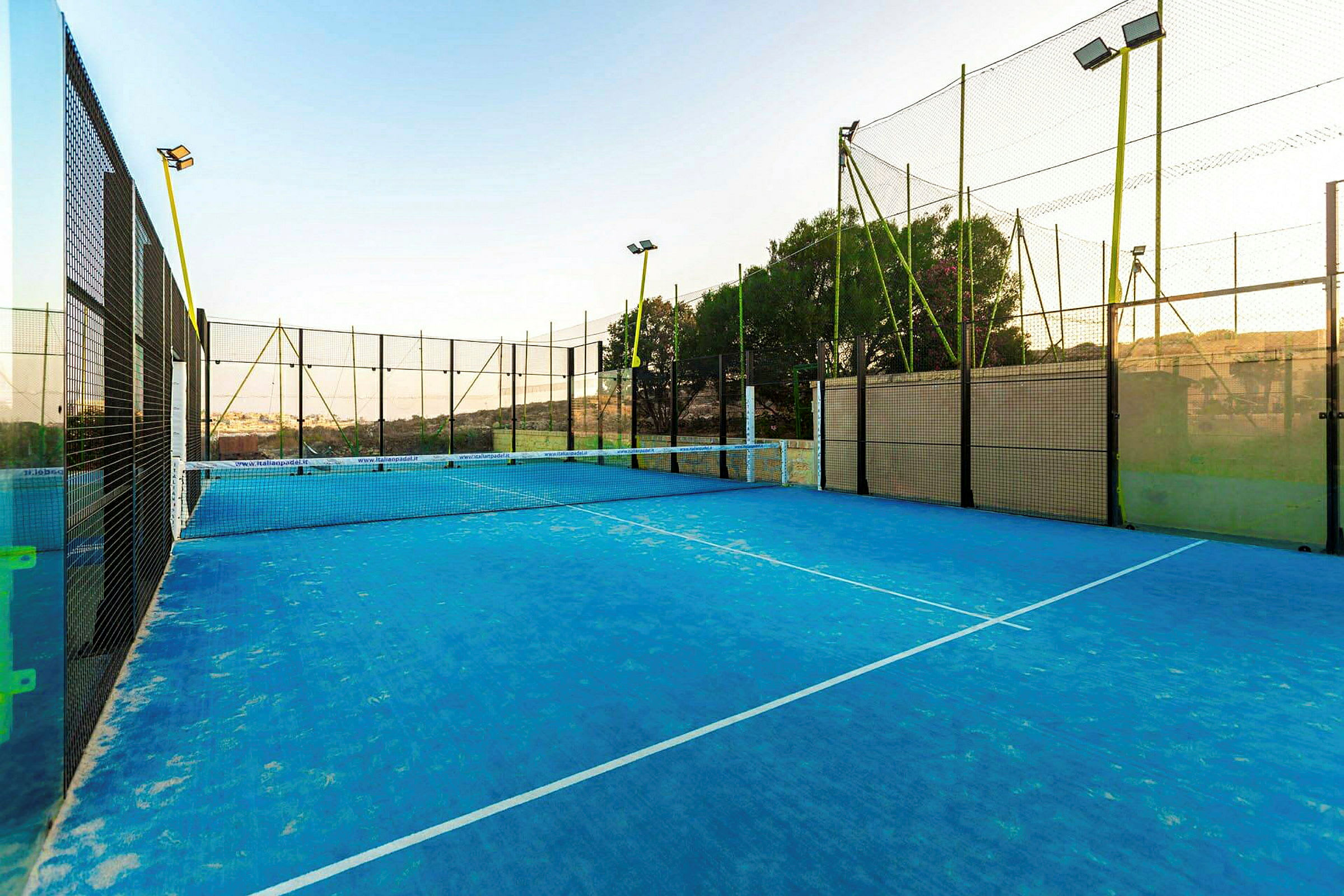 En padelbana med blått underlag och omgiven av höga nät, under en klar himmel. (AI-genererad text)