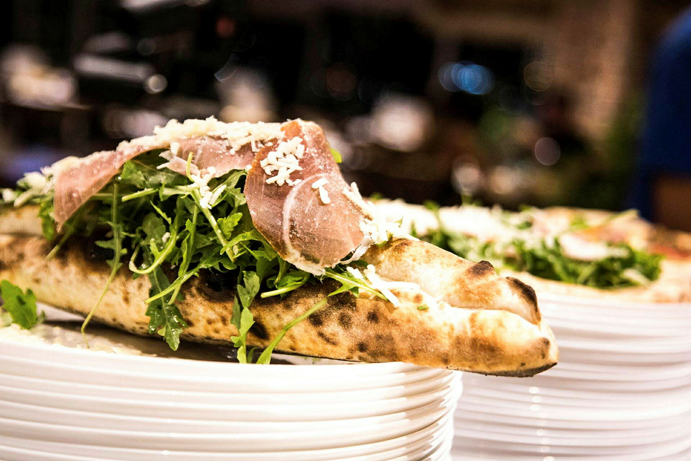 En närbild på en pizza med ruccolasallad och skinka, toppad med riven ost, serverad på en tallrik. (AI-genererad text)