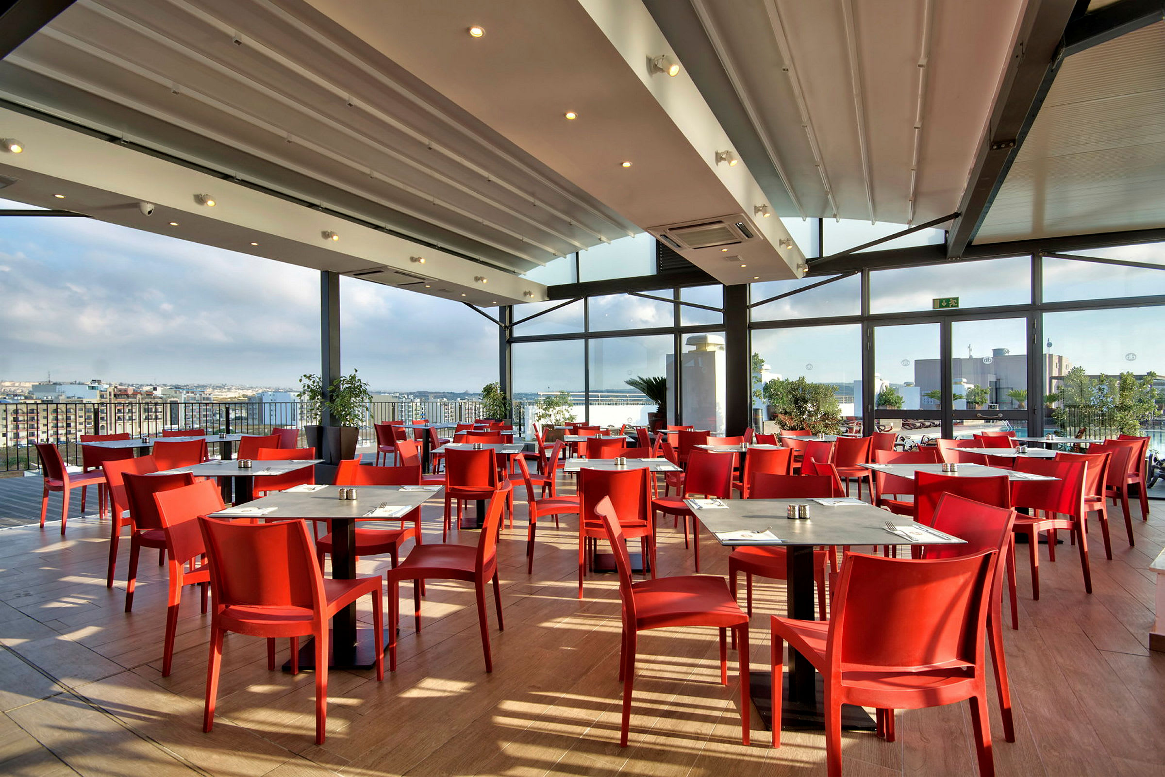 En ljus restaurang med röda stolar, träbord och stora fönster med utsikt över en stad. (AI-genererad text)