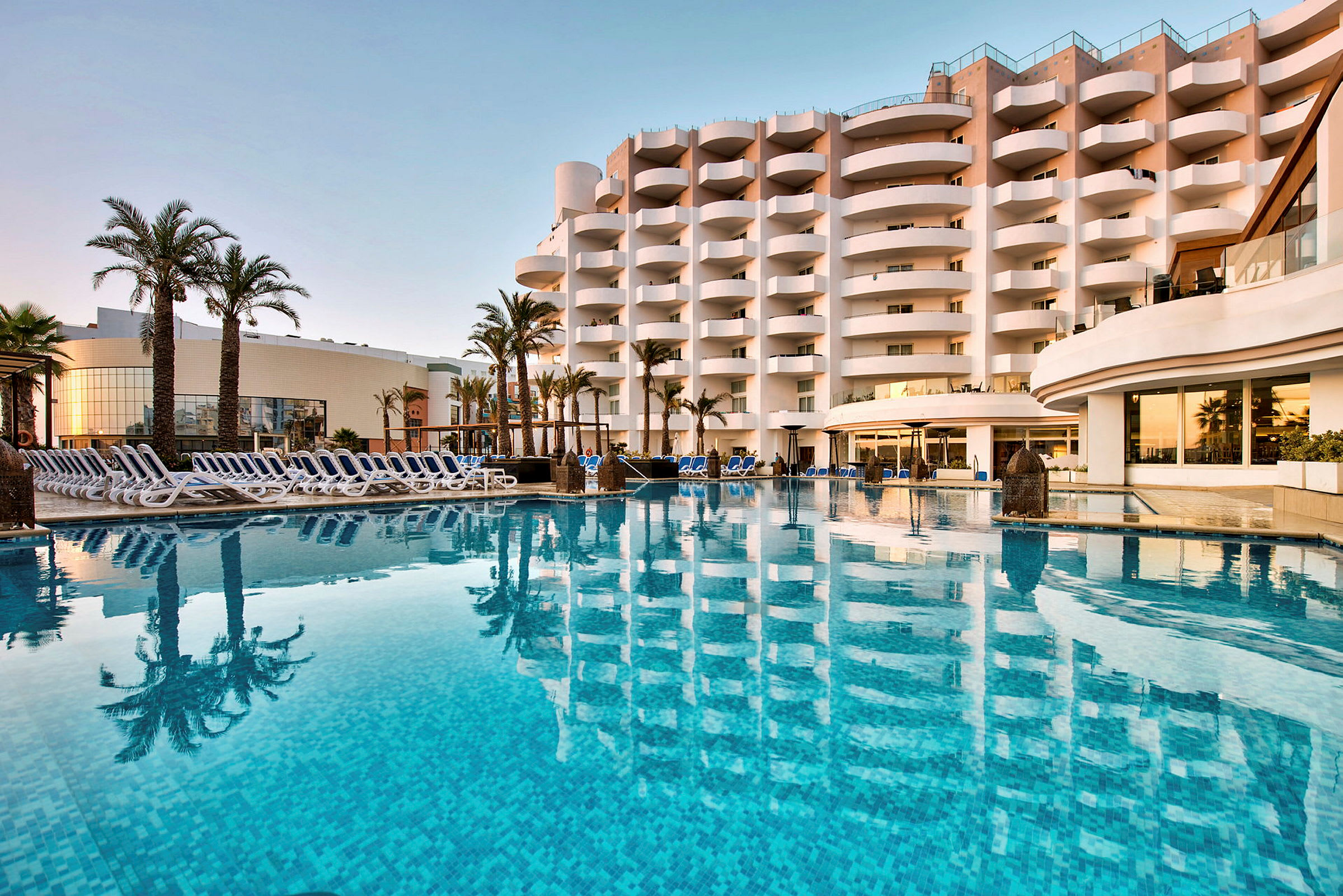Hotellbyggnad vid en utomhuspool med stolar och palmer. (AI-genererad text)