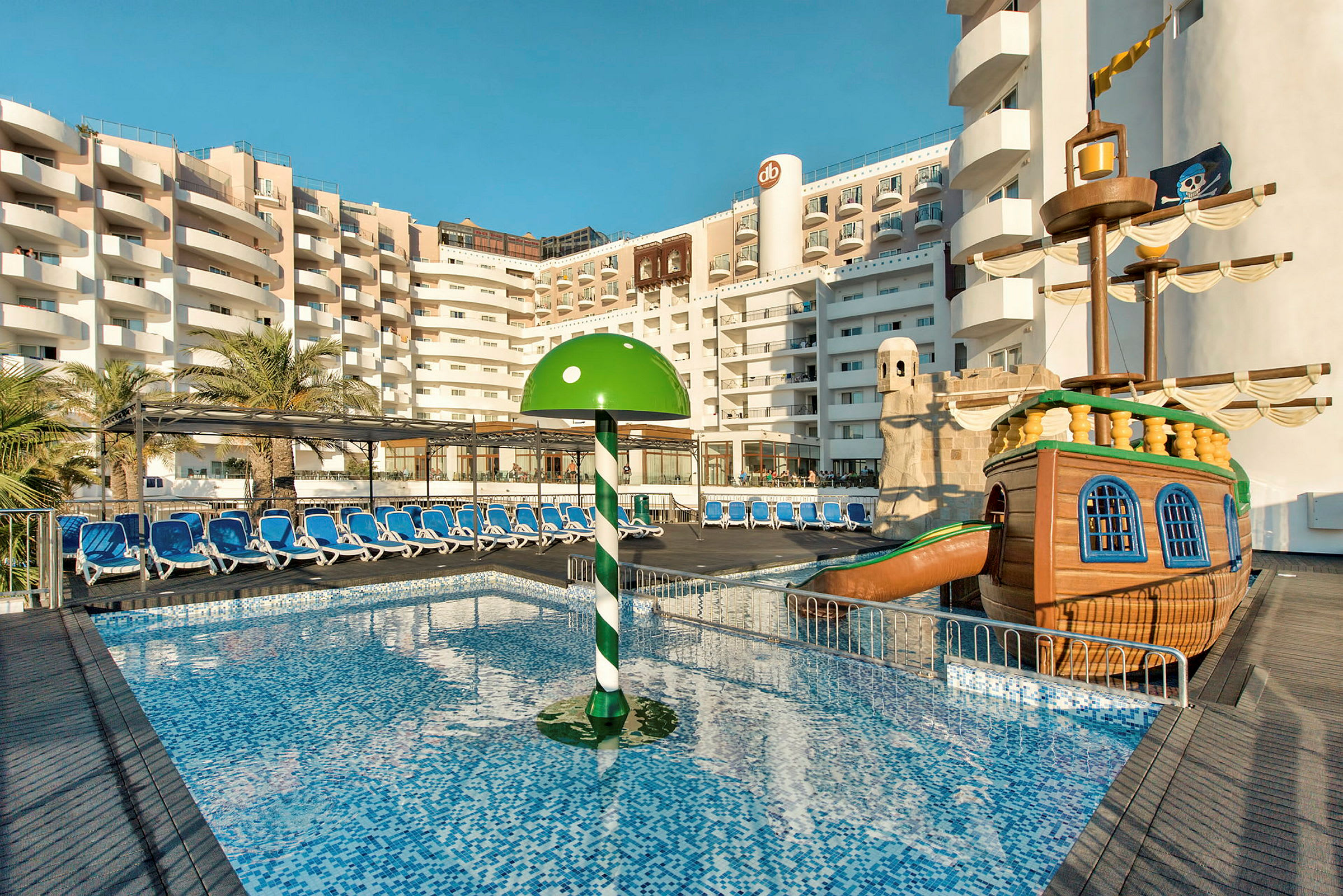 En hotellpool med solstolar, en grön parasoll och en lekpiratbåt. (AI-genererad text)