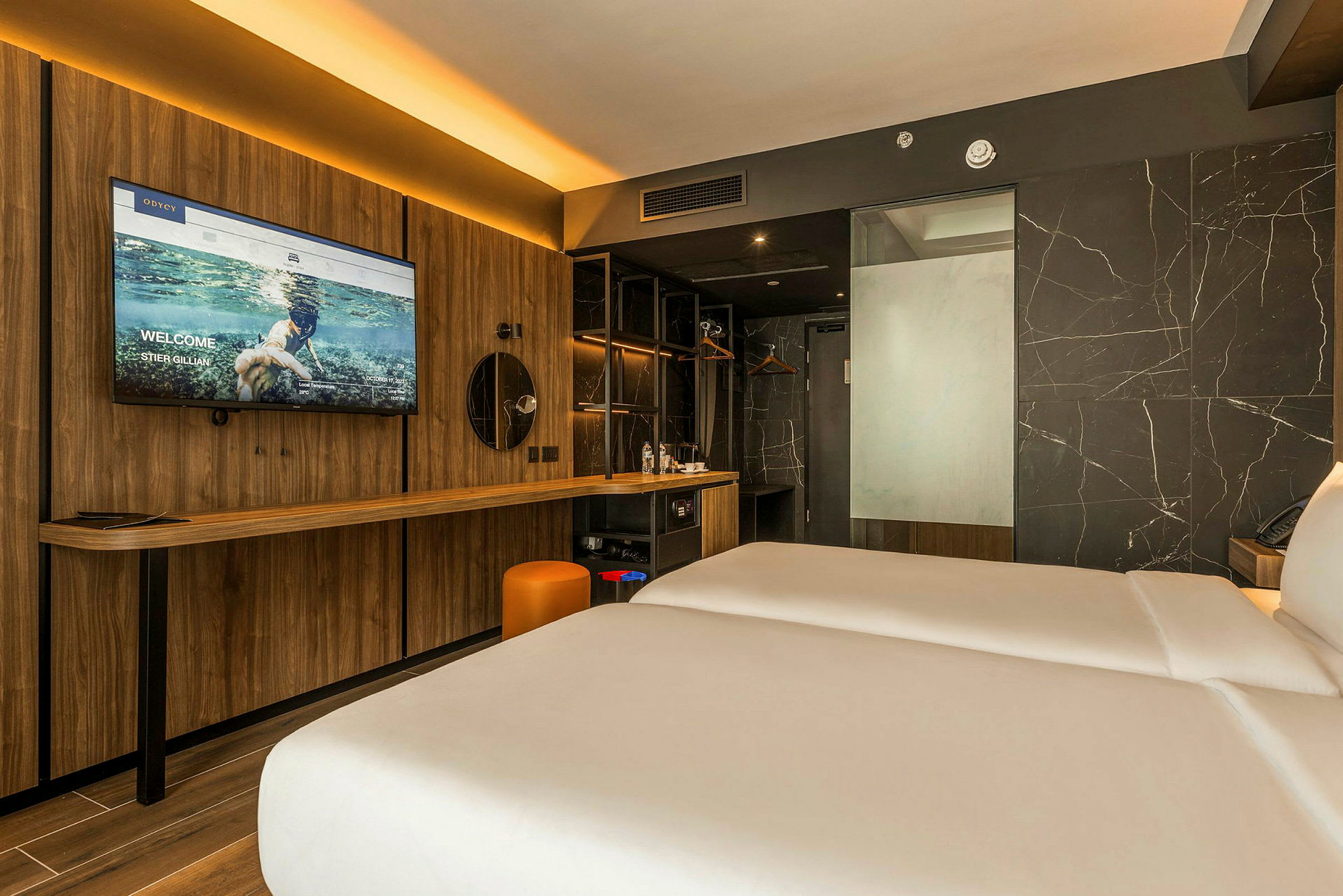 Ett modernt hotellrum med två sängar, en träpanel med en stor TV och en spegel, ett skrivbord samt en marmordekorerad vägg. (AI-genererad text)