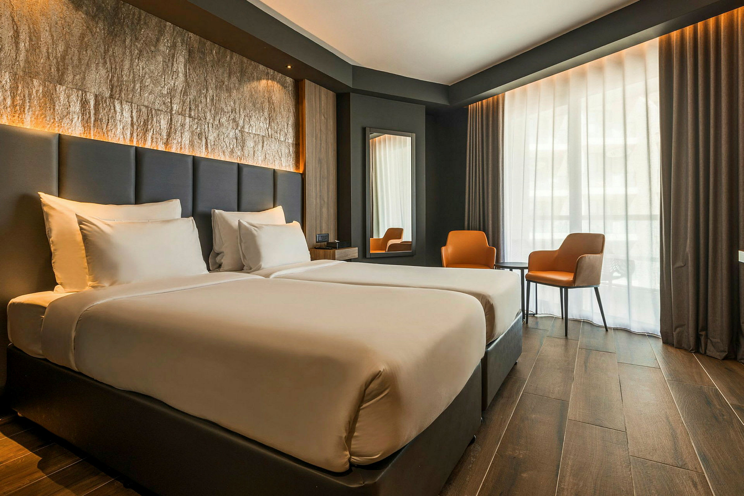 Ett hotellrum med två enkelsängar, en stor fönsteryta med gardiner, två oranga stolar och ett bord. (AI-genererad text)