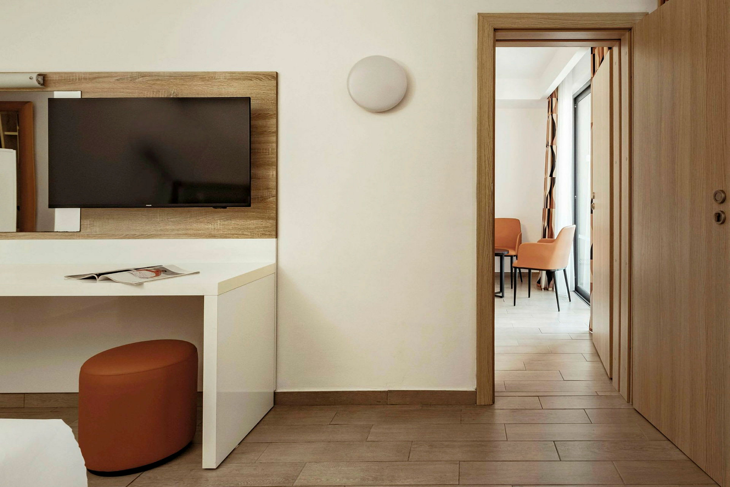Ett hotellrum med en TV på väggen ovanför ett vitt skrivbord. En dörr öppnar till ett område med två stolar och ett bord. (AI-genererad text)