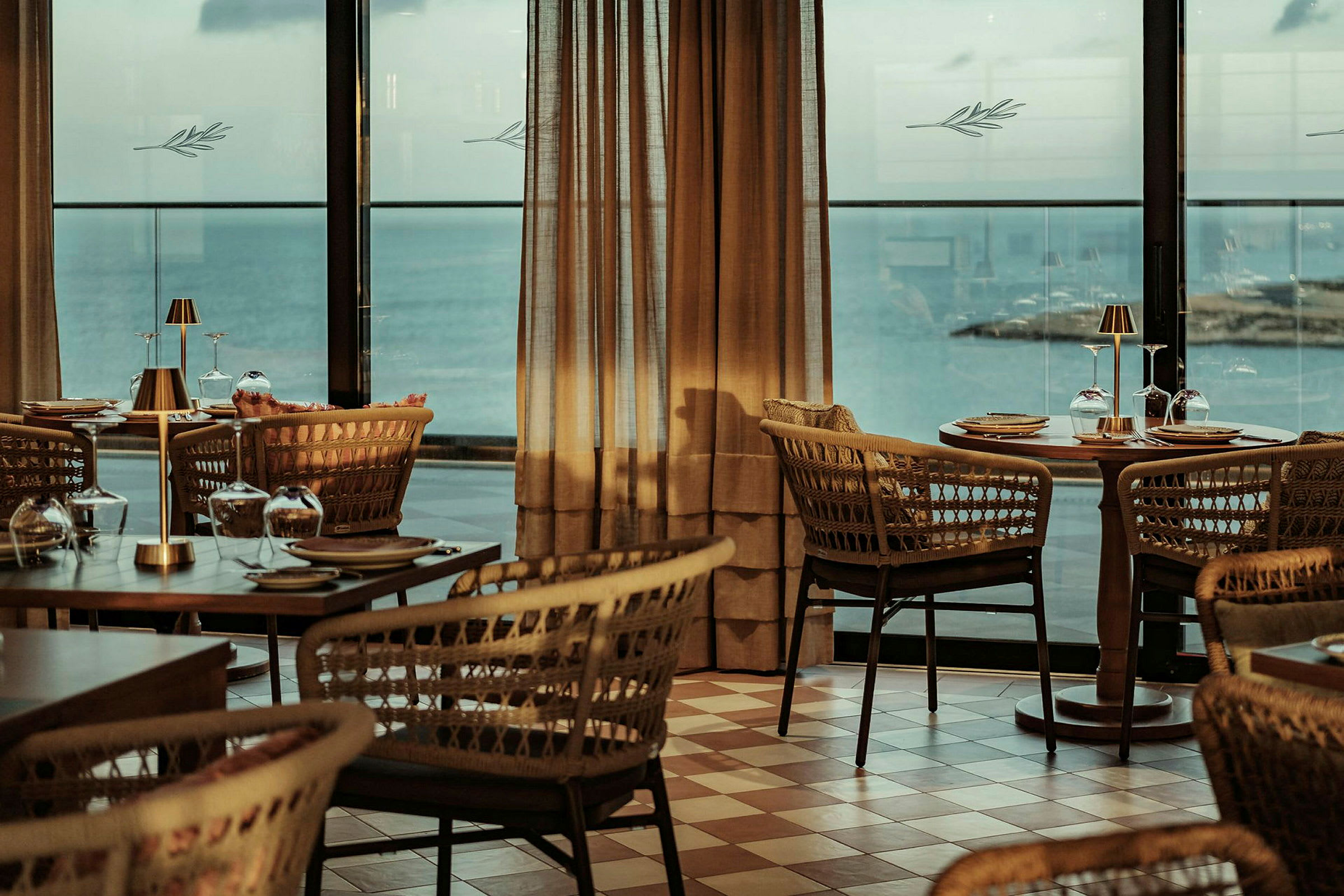 En restaurang med flätade stolar och dukade träbord med utsikt över havet genom stora fönster. (AI-genererad text)