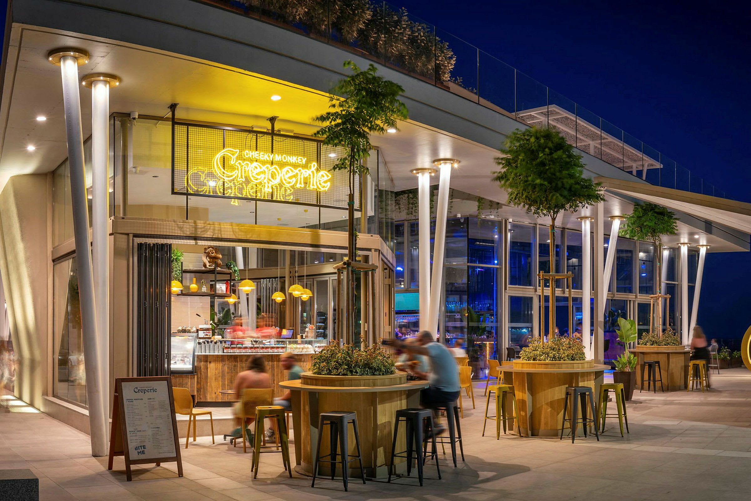 En modern restaurang eller café med skyltning som lyser, sittplatser utomhus och en väl upplyst fasad på kvällen. Träd och växtdekorationer omger områdena. (AI-genererad text)