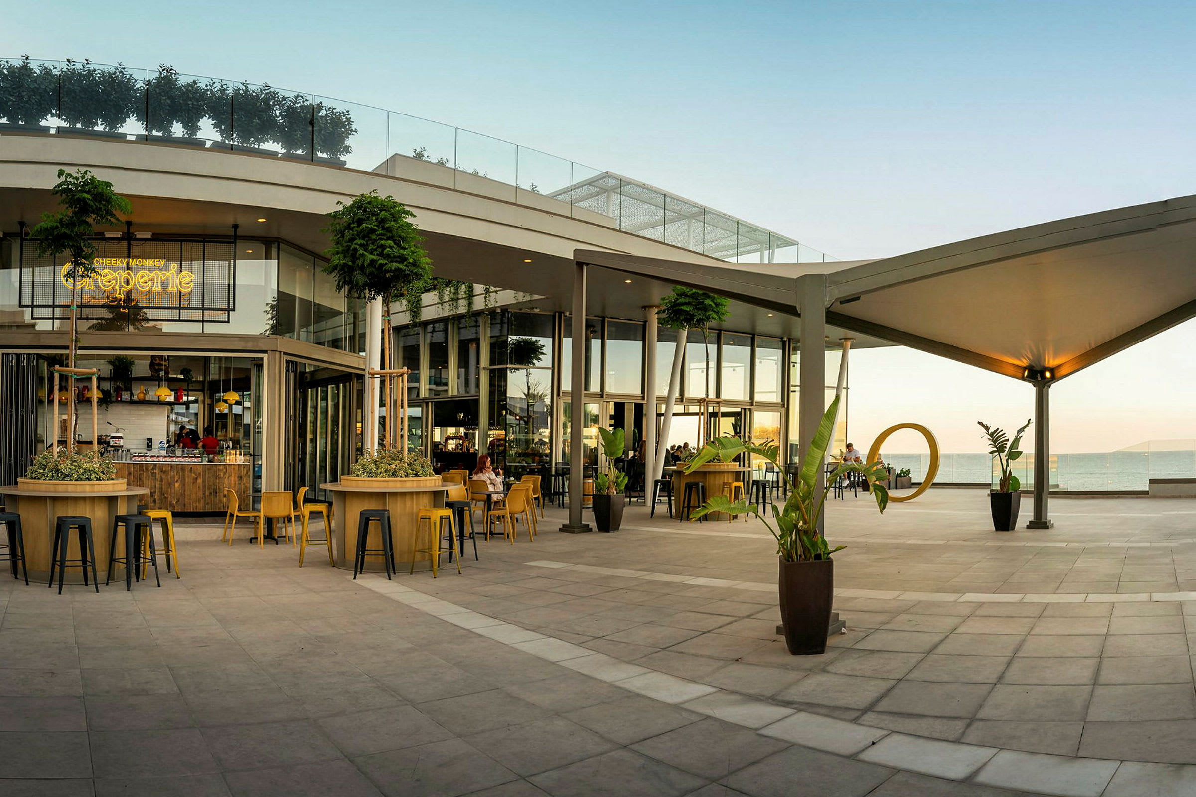 En modern utomhusterrass med sittplatser och planteringar vid en cafébyggnad mot havet. (AI-genererad text)
