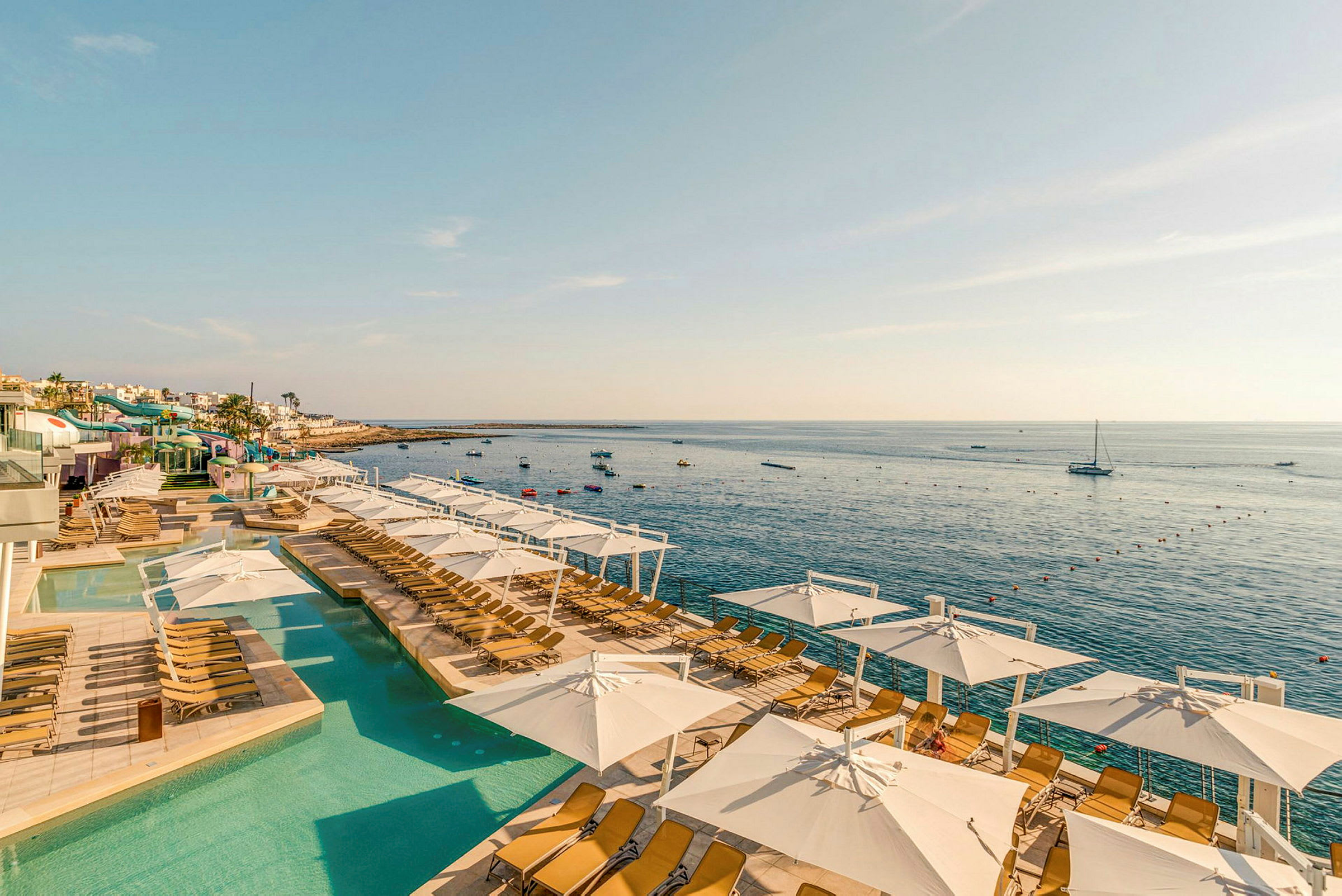 Utsikt över en pool med solstolar och parasoller nära havet, med små båtar som flyter i vattnet. (AI-genererad text)