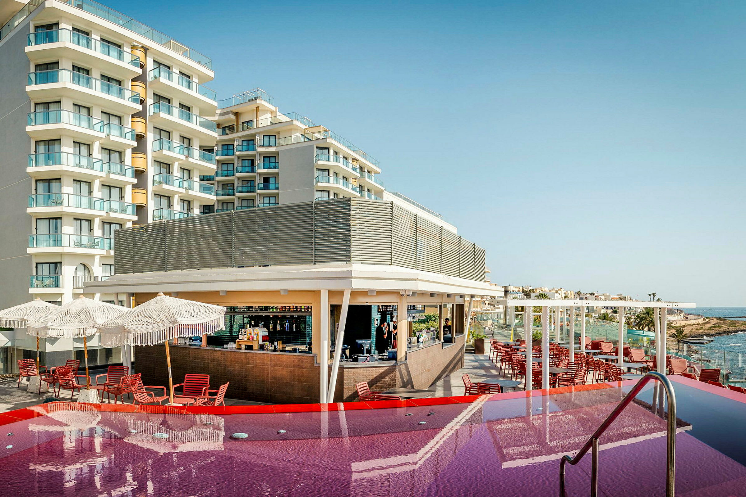Ett hotell med balkonger och en pool framför, samt en bar och sittplatser under parasoller. (AI-genererad text)