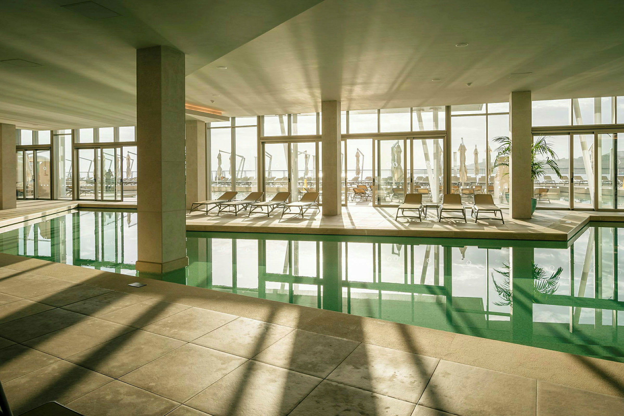 En inomhuspool i ett ljusfyllt rum med stora fönster som vetter mot en terrass med solstolar. (AI-genererad text)