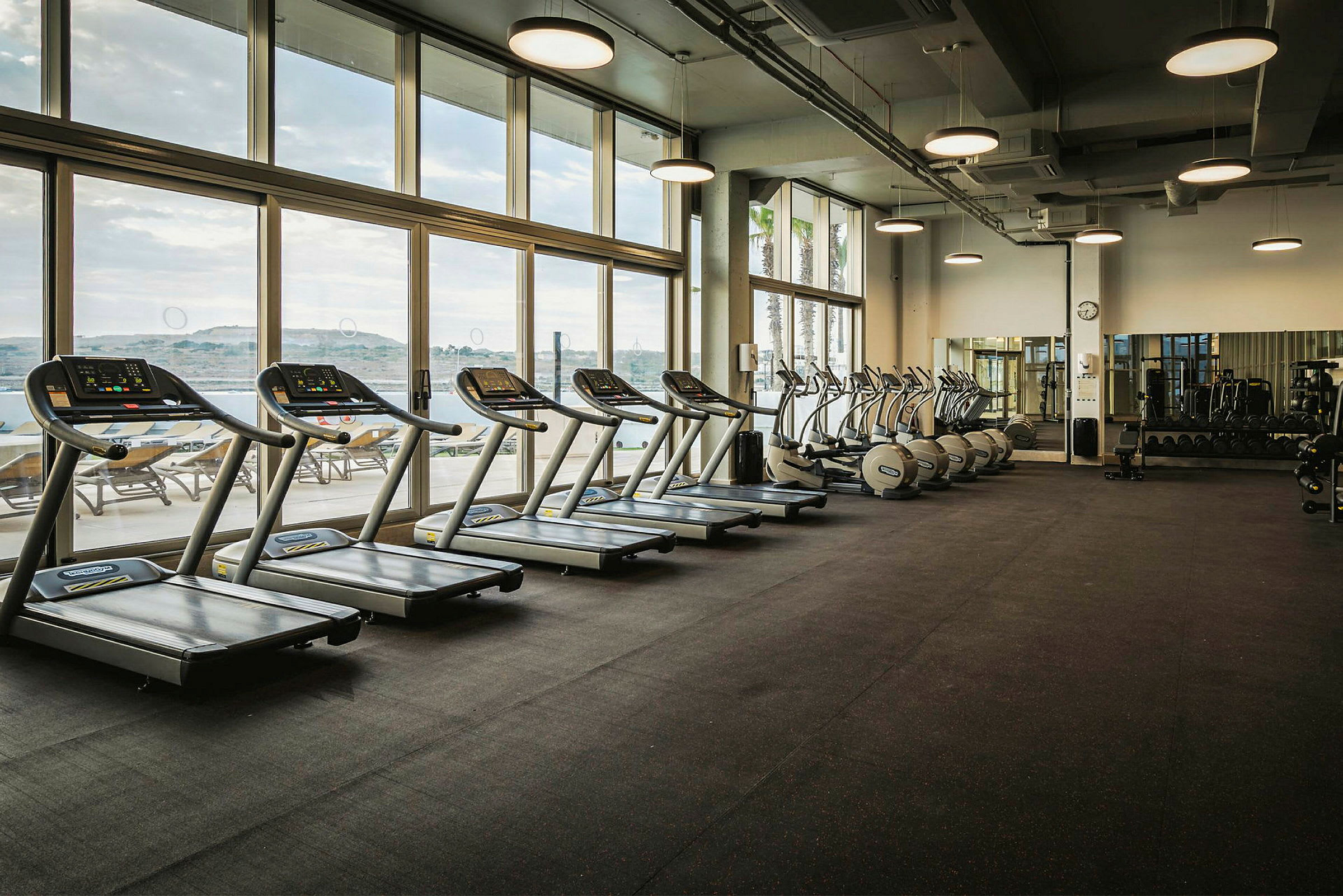 Ett gym med träningsmaskiner, inklusive löpband, motionscyklar och vikter, med stora fönster som släpper in dagsljus. (AI-genererad text)