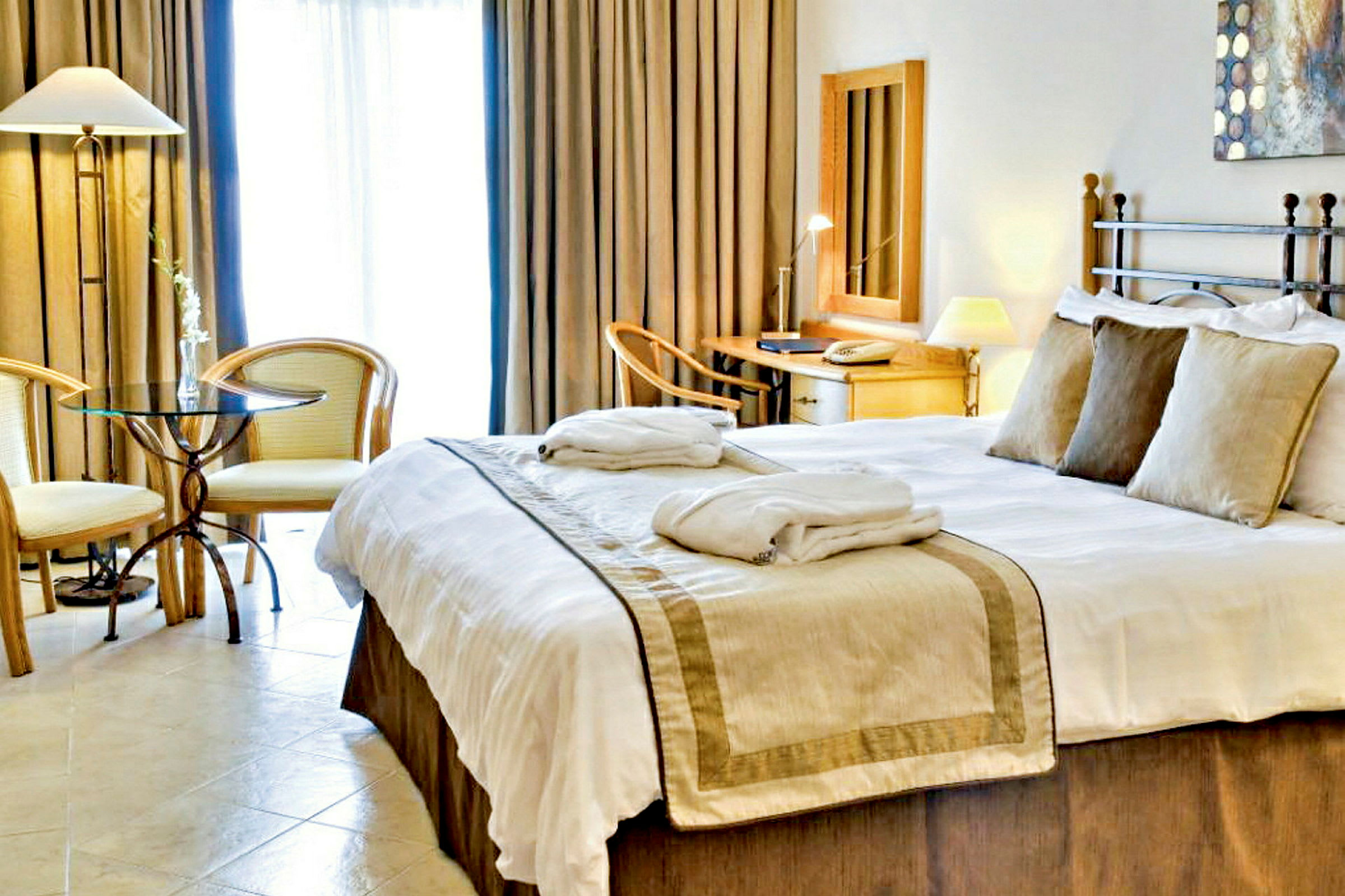 Ett hotellrum med en stor säng, sängkläder och kuddar. Två fåtöljer och ett bord står vid sidan, och en arbetsplats med spegel och skrivbord syns i bakgrunden. (AI-genererad text)