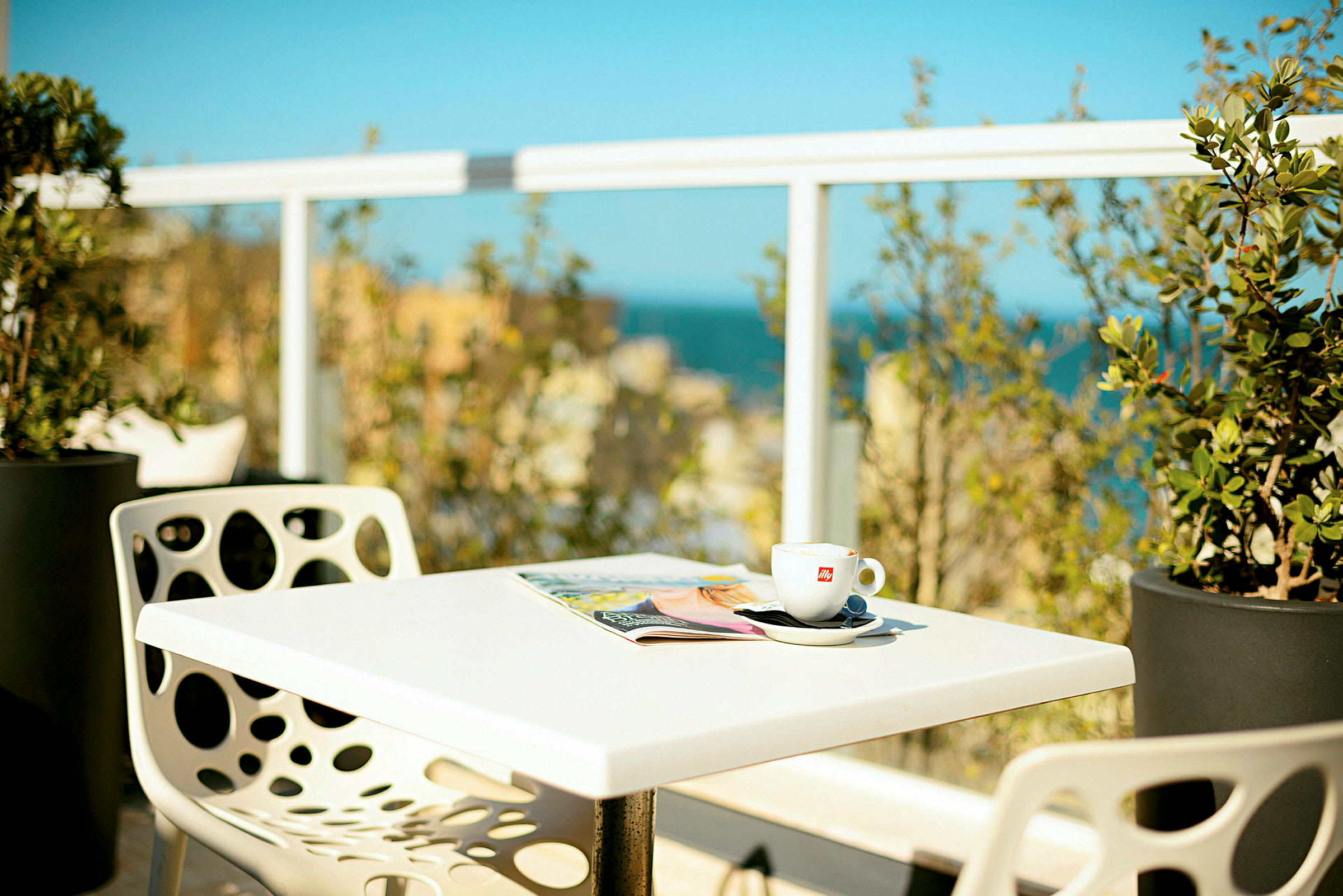 Ett bord med en kaffekopp och en tidning, omgivet av grönska och med utsikt över havet i bakgrunden. (AI-genererad text)