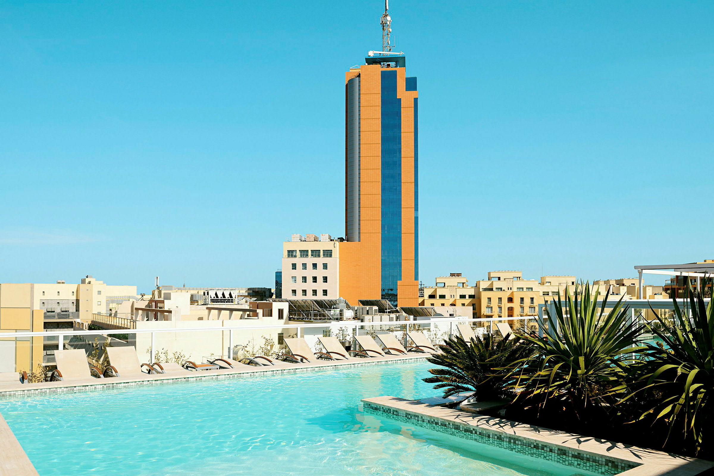 Bild av ett hotellområde med pool, solstolar och grönska i förgrunden. En hög byggnad omgiven av lägre byggnader syns i bakgrunden. (AI-genererad text)