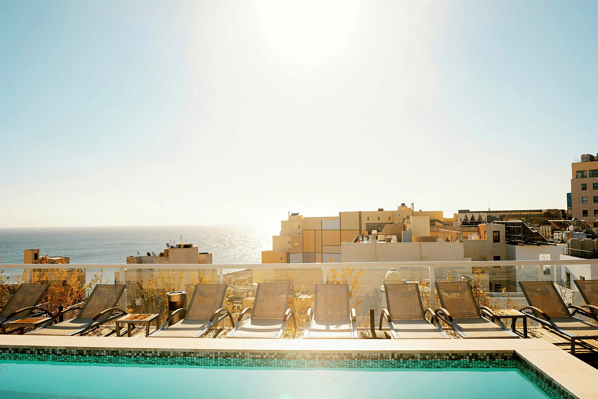 En pool med solstolar framför en utsikt mot havet och byggnader. (AI-genererad text)
