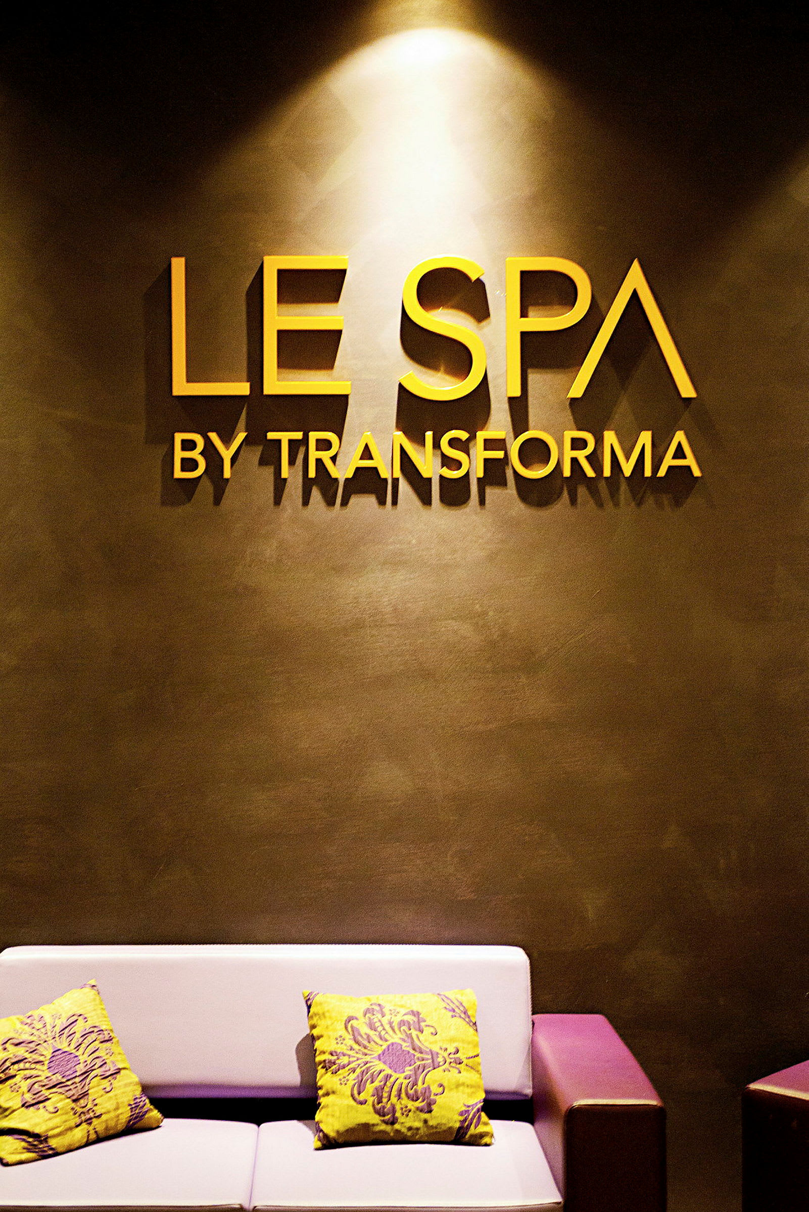 Bild av en spa-entré med texten "Le Spa by Transforma" på väggen. En soffa med färgglada prydnadskuddar är synlig. (AI-genererad text)
