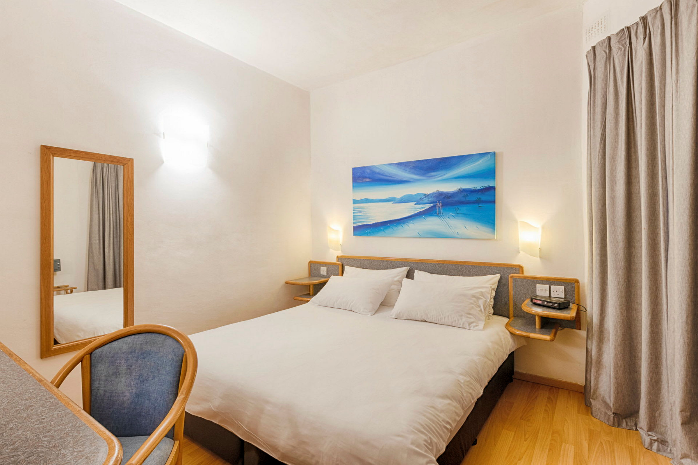 Ett hotellrum med en dubbelsäng, ett skrivbord med en stol och en spegel. Rummet har ljusa väggar och en väggdekoration med strandmotiv. (AI-genererad text)